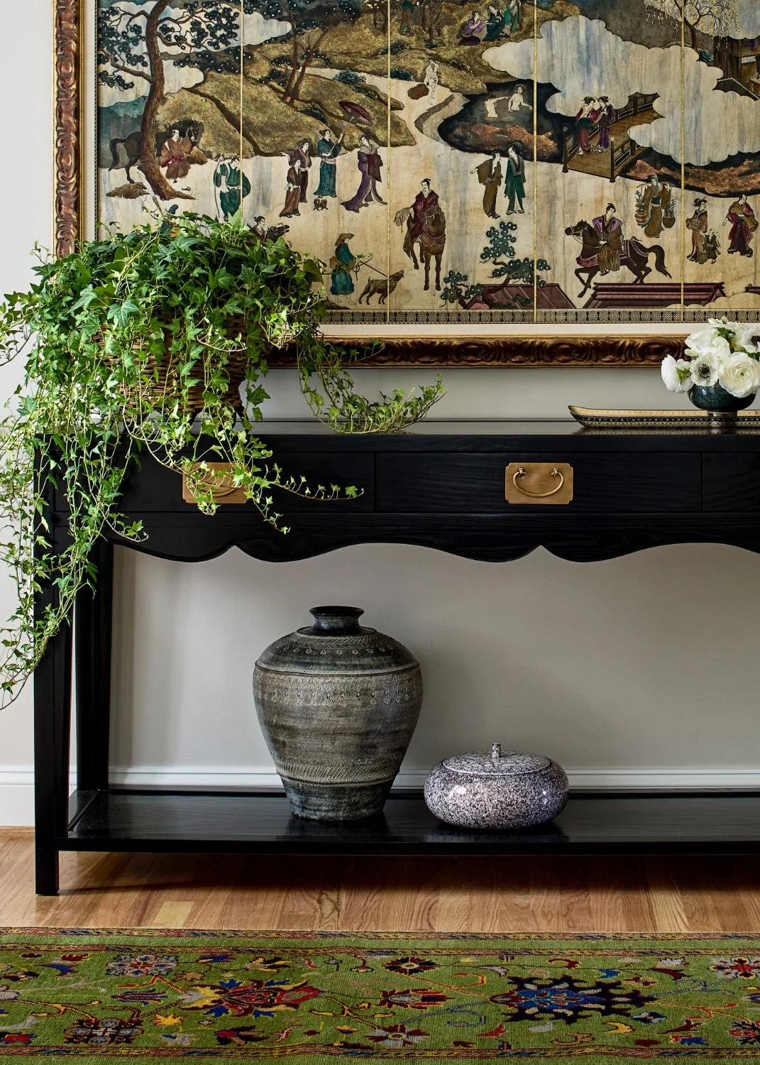 Interior with black console table, large potted ivy on the left, decorative jars on the shelf, artwork of a traditional Asian scene with people and nature, floral arrangement in a dark vase, colorful patterned rug on a wooden floor.