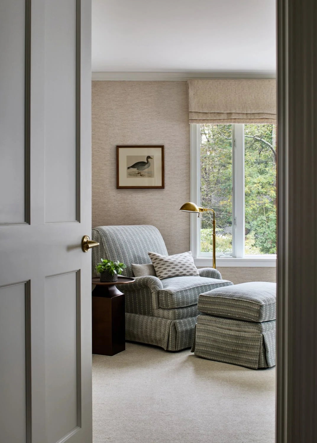 View of a cozy living room with a striped armchair, a matching ottoman, a side table with a potted plant, a gold floor lamp, a framed duck print on the textured wall, and a window with a view of trees outside.