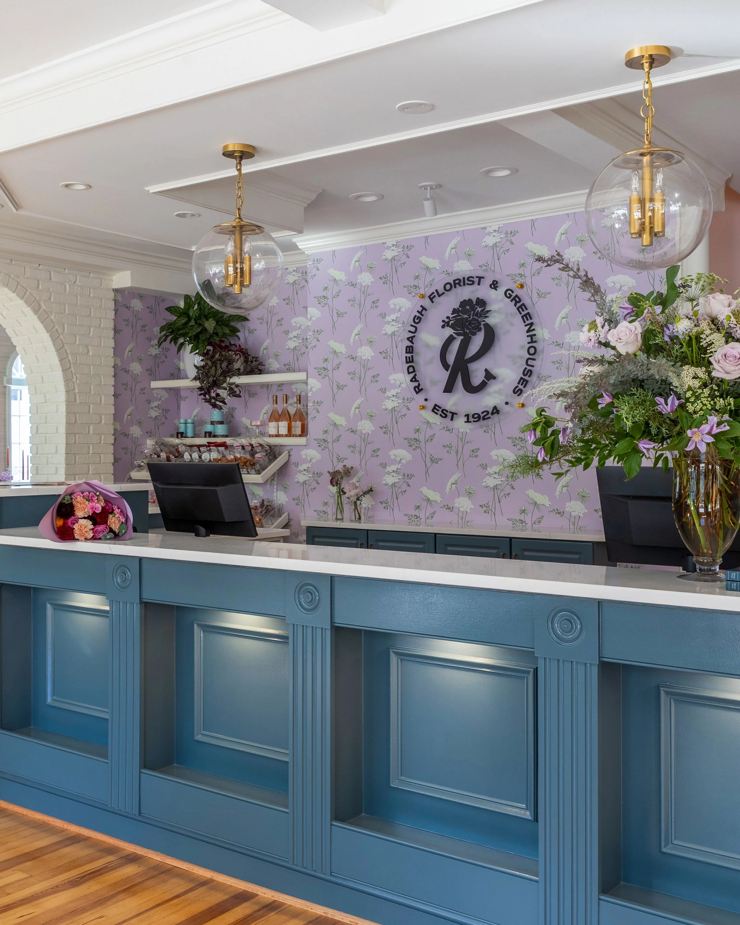 Interior of a florist shop with a blue counter, floral arrangements, and a purple floral wallpaper wall with the business logo and name.