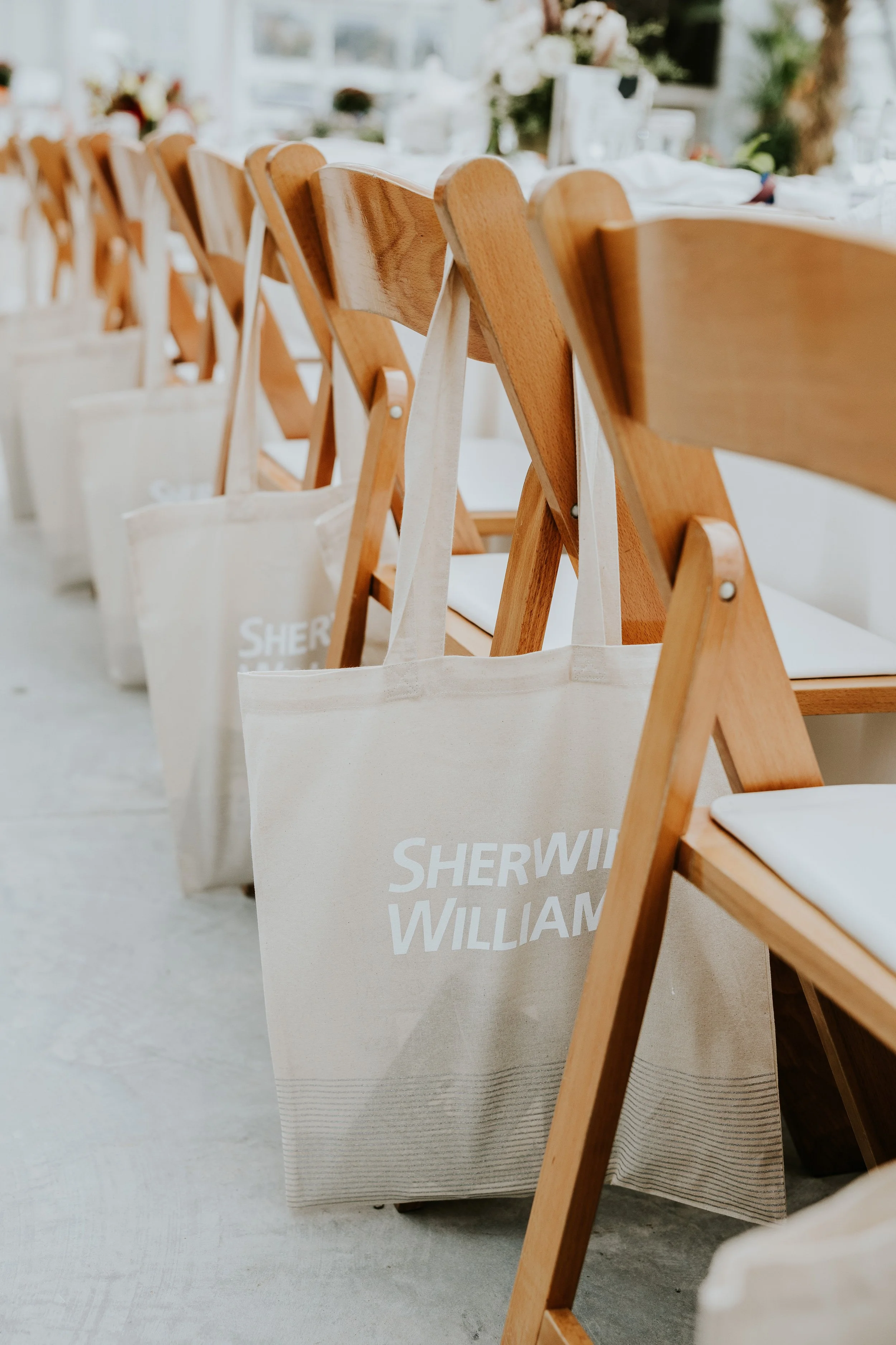 Line of wooden chairs with white fabric tote bags hanging on their backs, placed along a table set for an event or gathering.