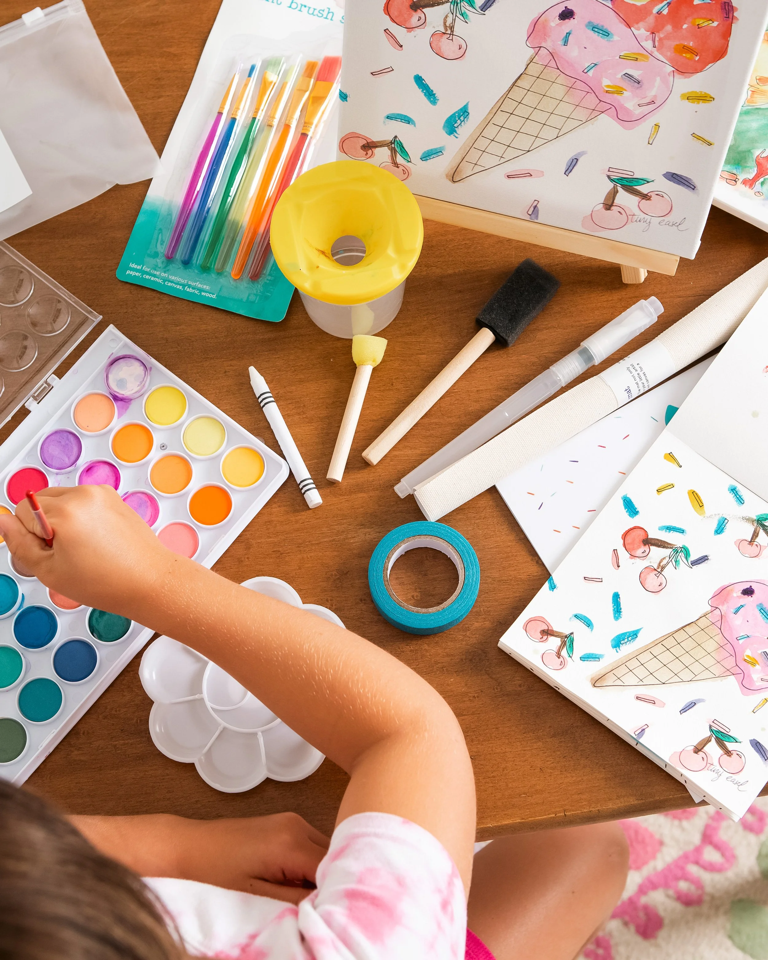 Child painting with watercolor paints on a table with art supplies, including paint palettes, brushes, washi tape, and drawings of ice cream and cherries.