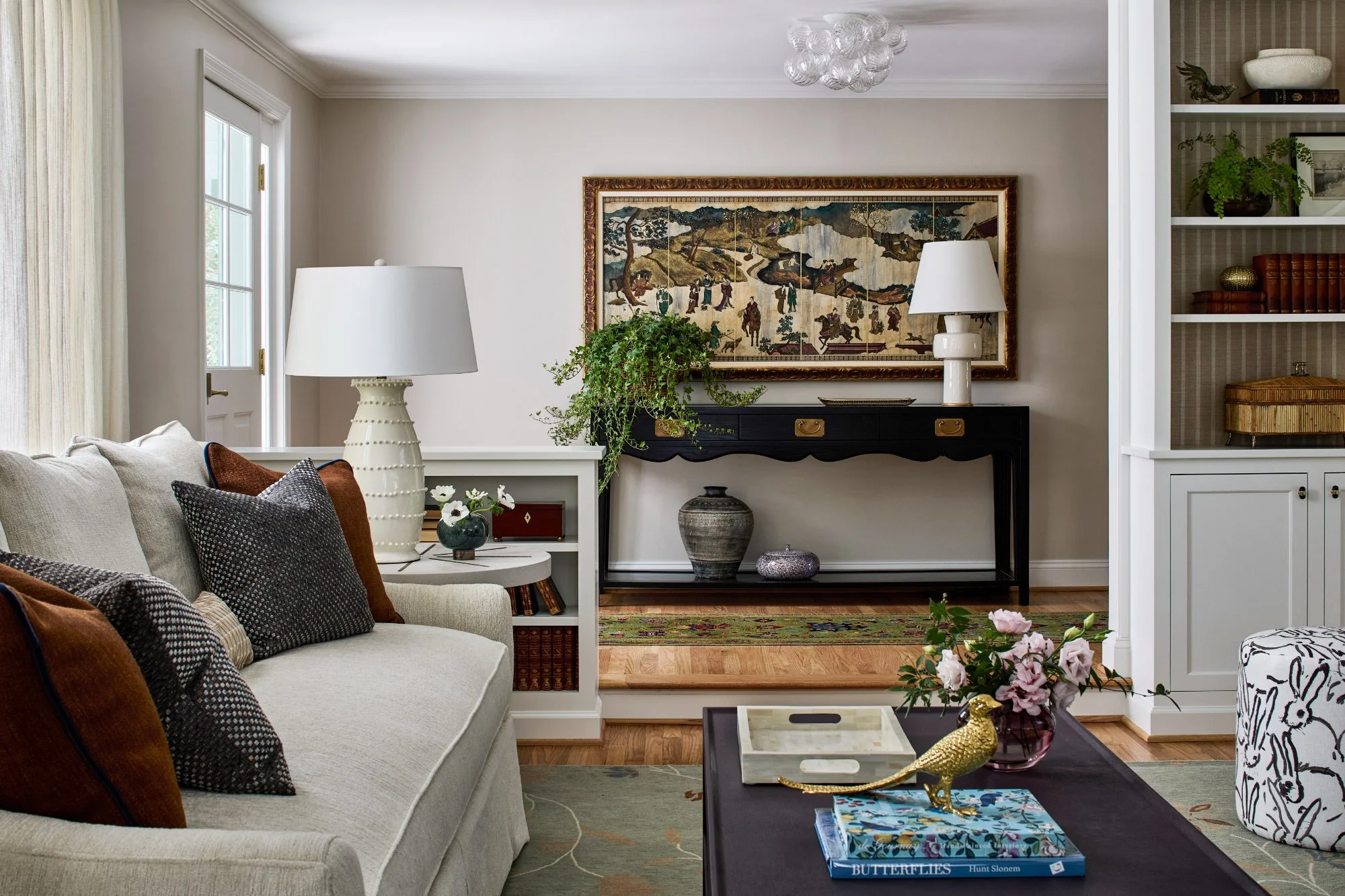 A living room with a white sofa holding brown and black decorative pillows, a white side table with a lamp and small plant, a black console table with a large painting above, and a bookshelf with decorations and books. There is a coffee table with a book, a vase of pink flowers, and a golden bird figurine. The room has hardwood floors and a chandelier overhead.