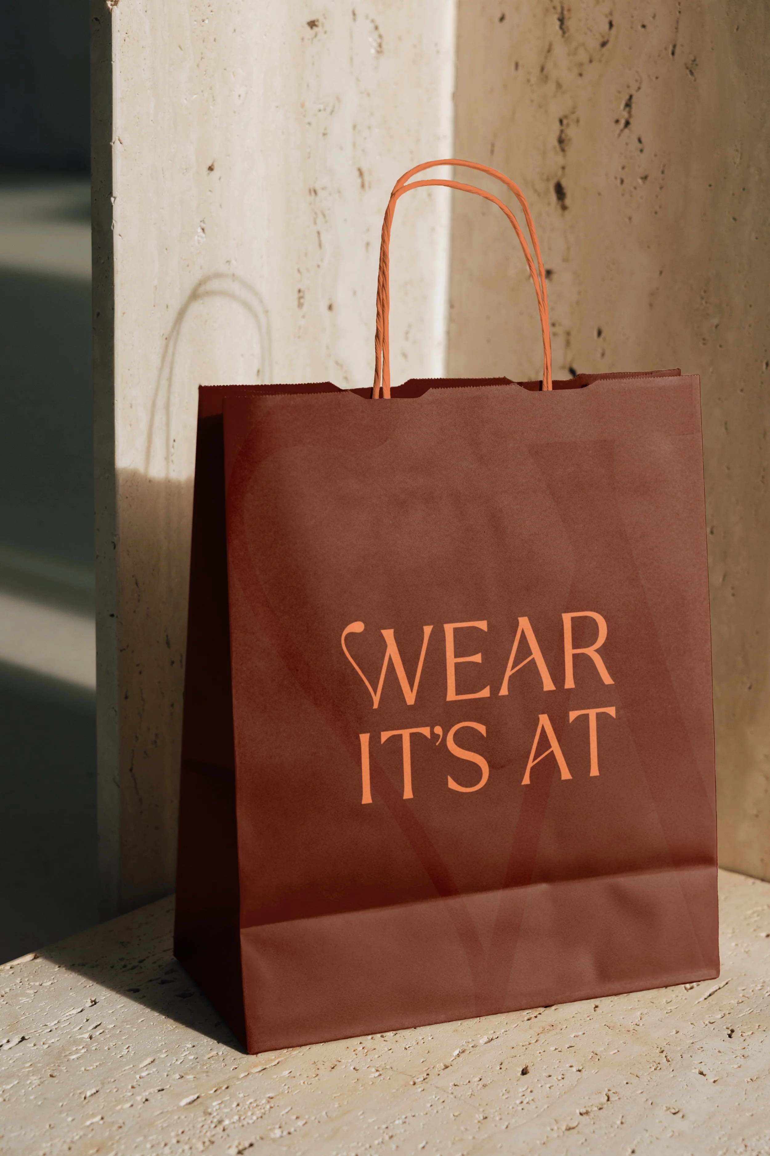 A pink shopping bag with orange handles, leaning against a stone wall, with the words "WEAR IT'S AT" printed on it.