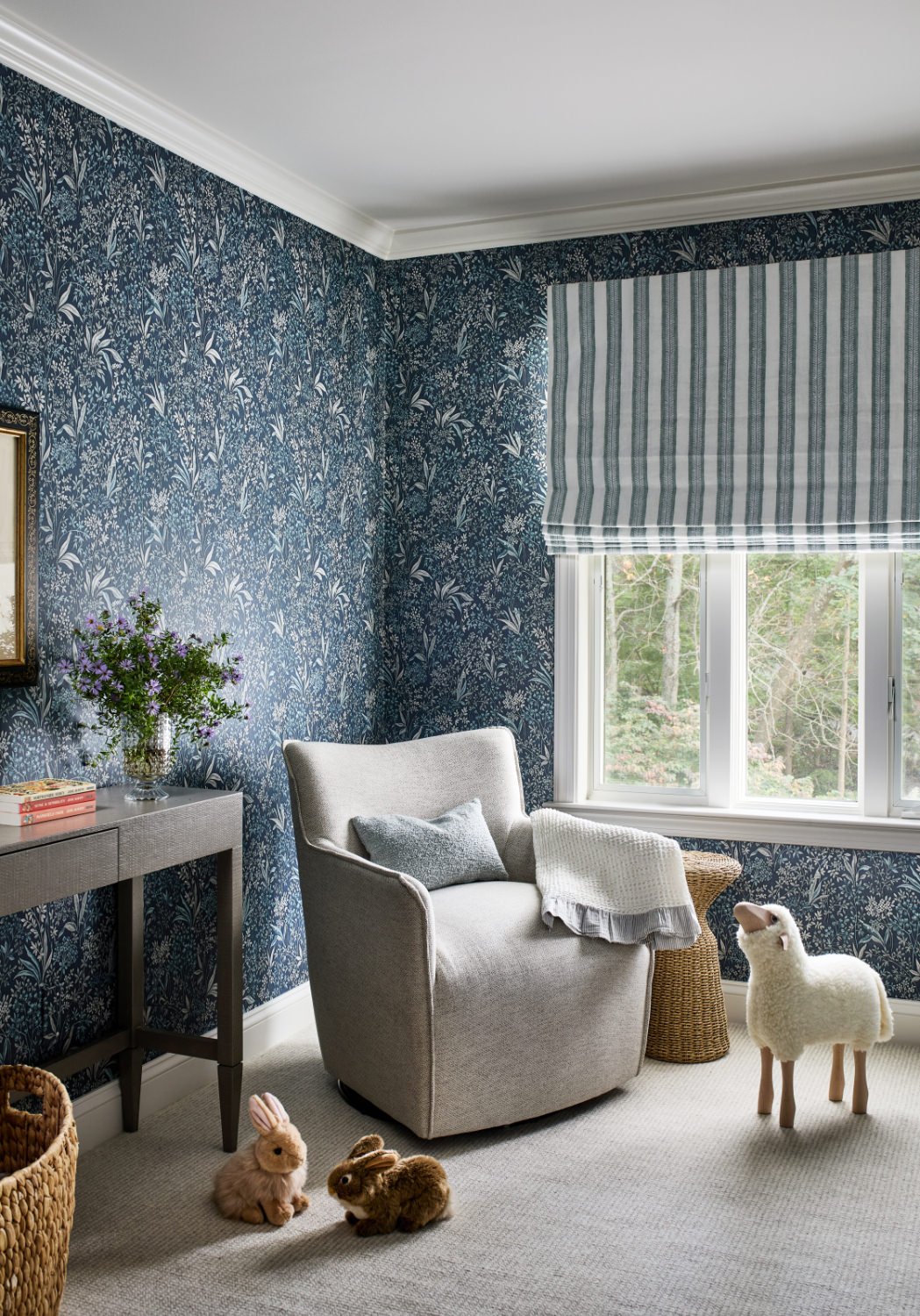 Interior of a cozy living room with a blue floral wallpaper, a large window with striped Roman shade, a beige armchair with gray pillow and throw, a wicker side table, a console table with a vase of purple flowers, and plush rabbit and sheep toys on a beige carpet.