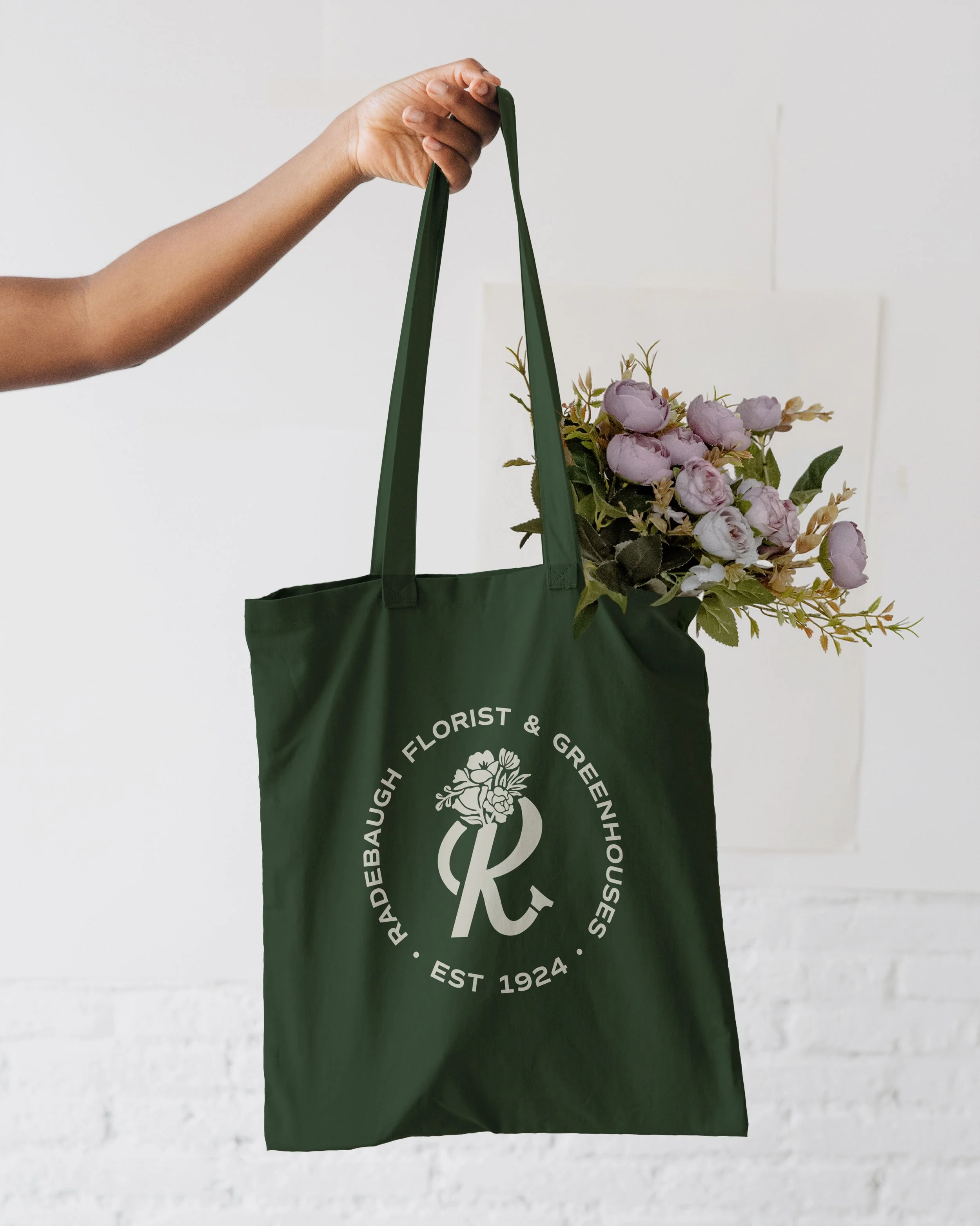 A person holding a green tote bag with floral logo by the handles, containing a bouquet of purple flowers, against a white wall background.