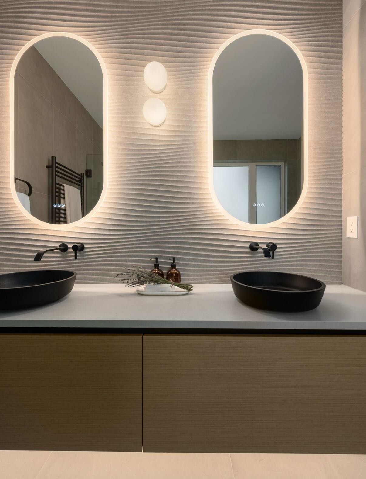 A bathroom that proves restraint can still feel bold.
Soft architectural curves, sculptural lighting, and a tactile feature wall set the tone, while matte black fixtures ground the space with contrast. Every detail was considered&mdash;down to the gl