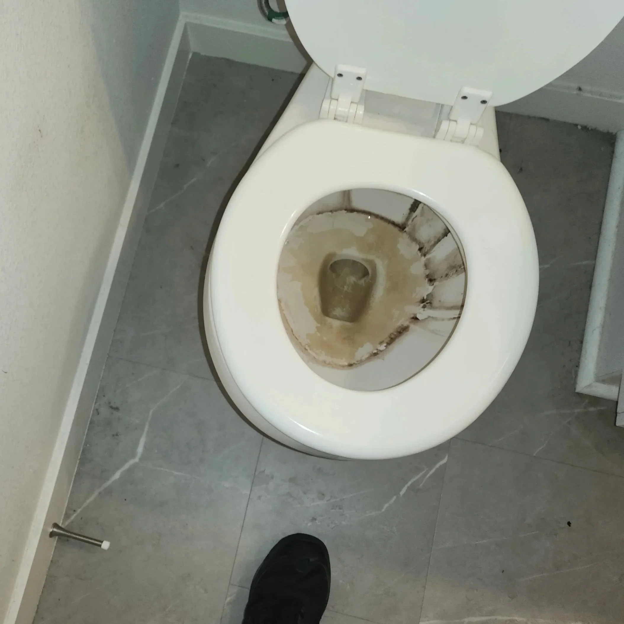 An empty toilet with a dirty bowl and open lid in a restroom with gray tiled floor and beige walls.