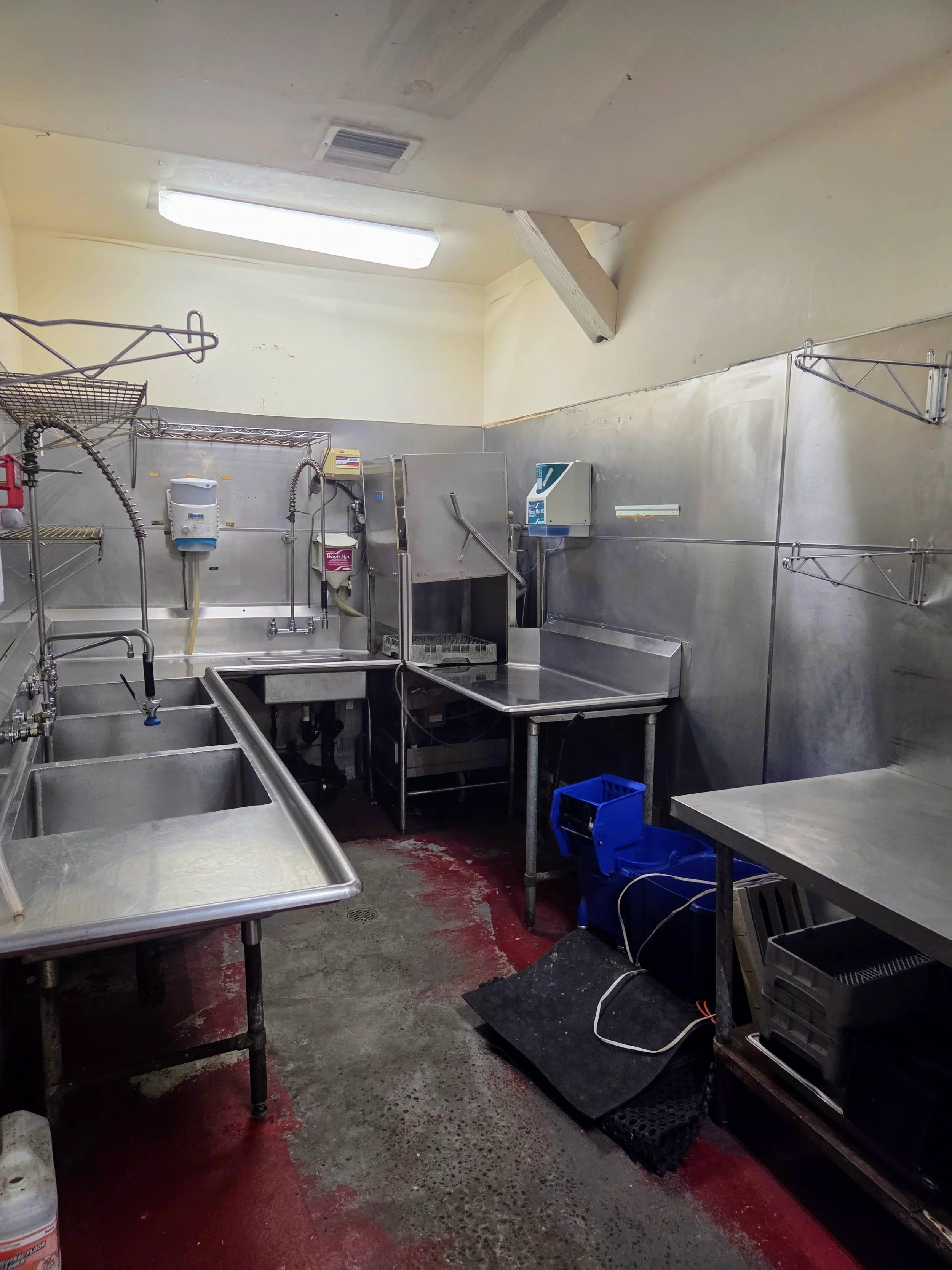 A commercial kitchen with stainless steel sinks, counters, shelves, and various cleaning supplies, featuring a concrete floor with red stains, fluorescent lighting, and a beige wall and ceiling.