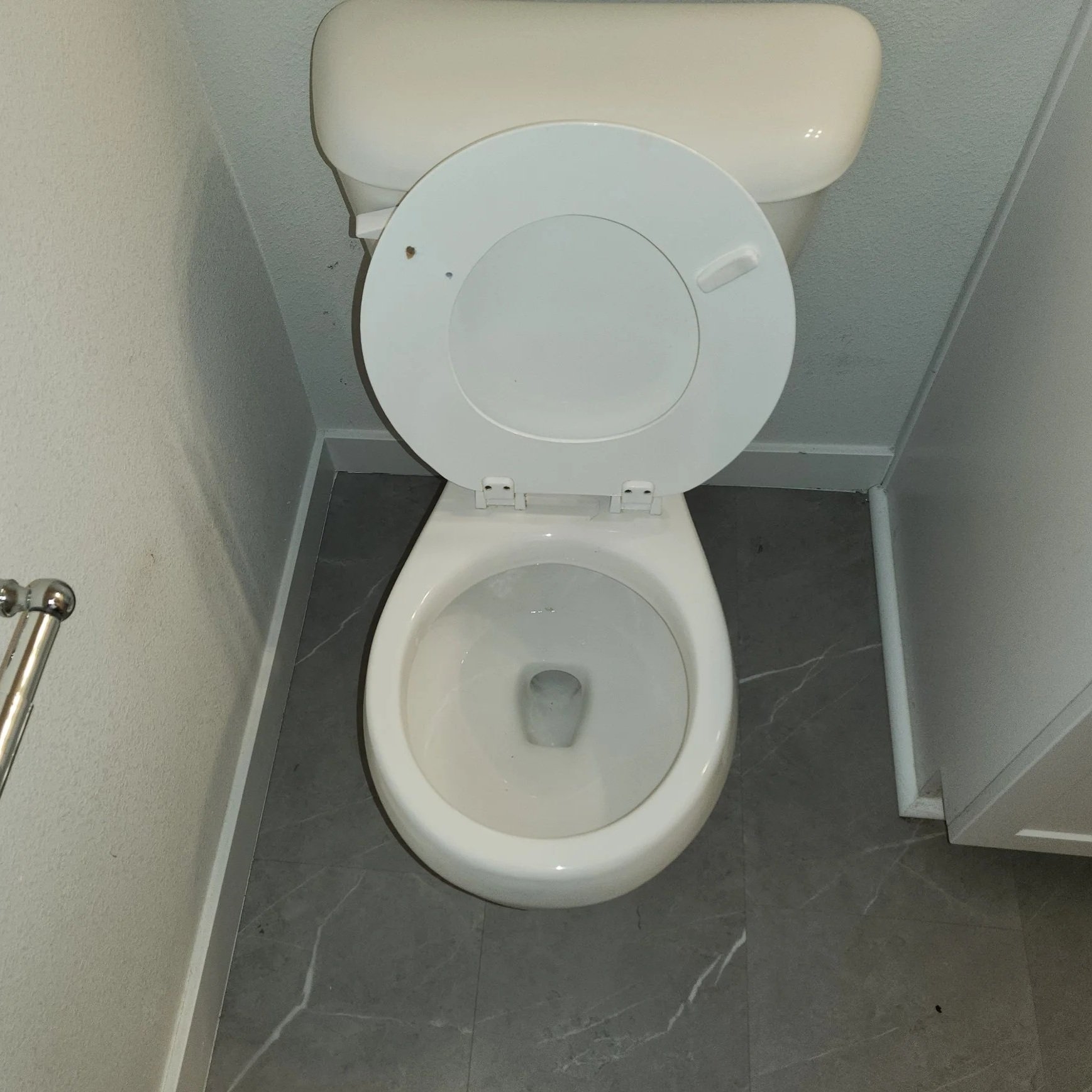 Unoccupied white toilet with its lid up in a bathroom with gray tiled flooring and beige wall.