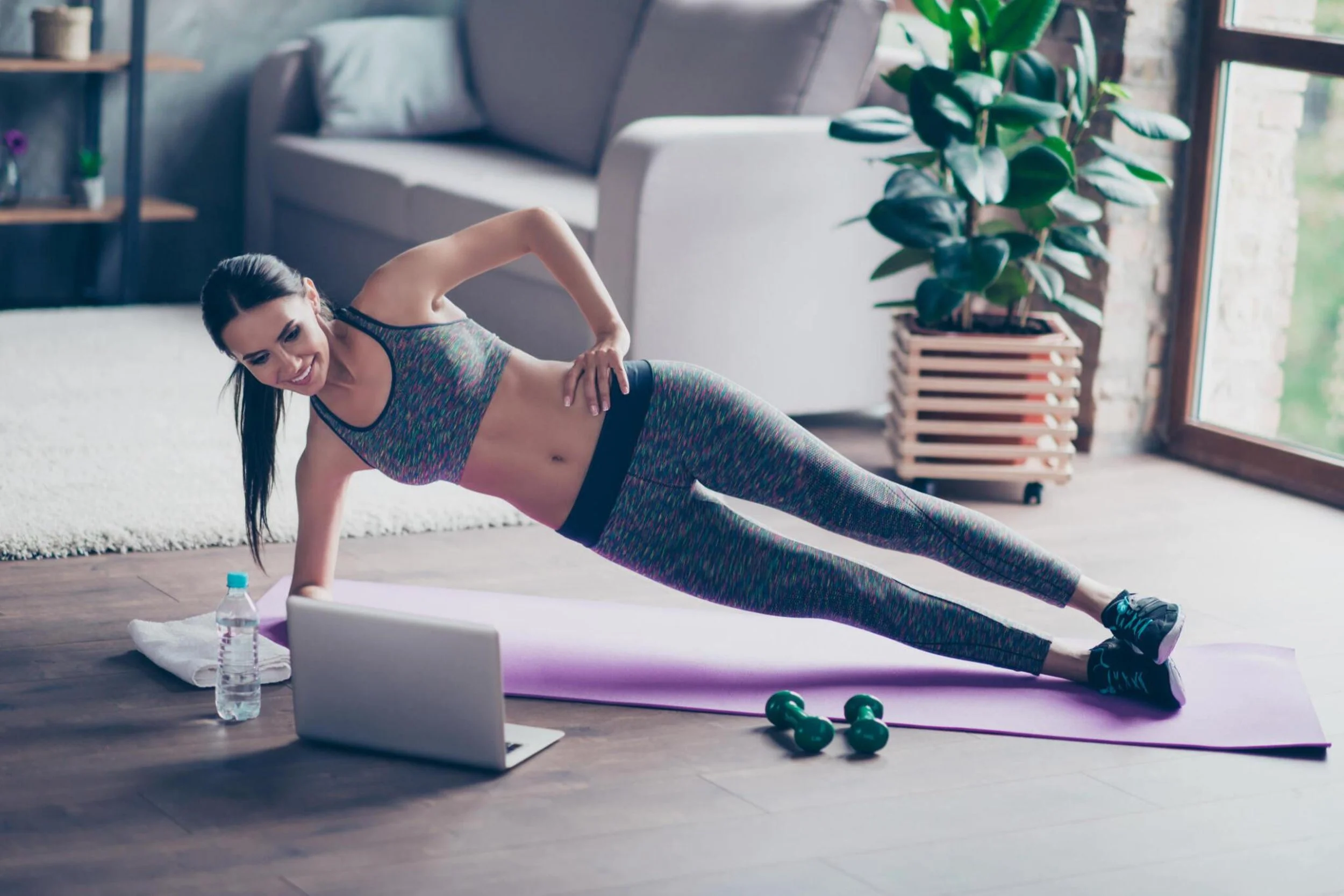 A woman doing an exercise or yoga move on a purple mat in a bright, airy living room with large windows, a gray sofa, indoor plants, and wooden flooring.