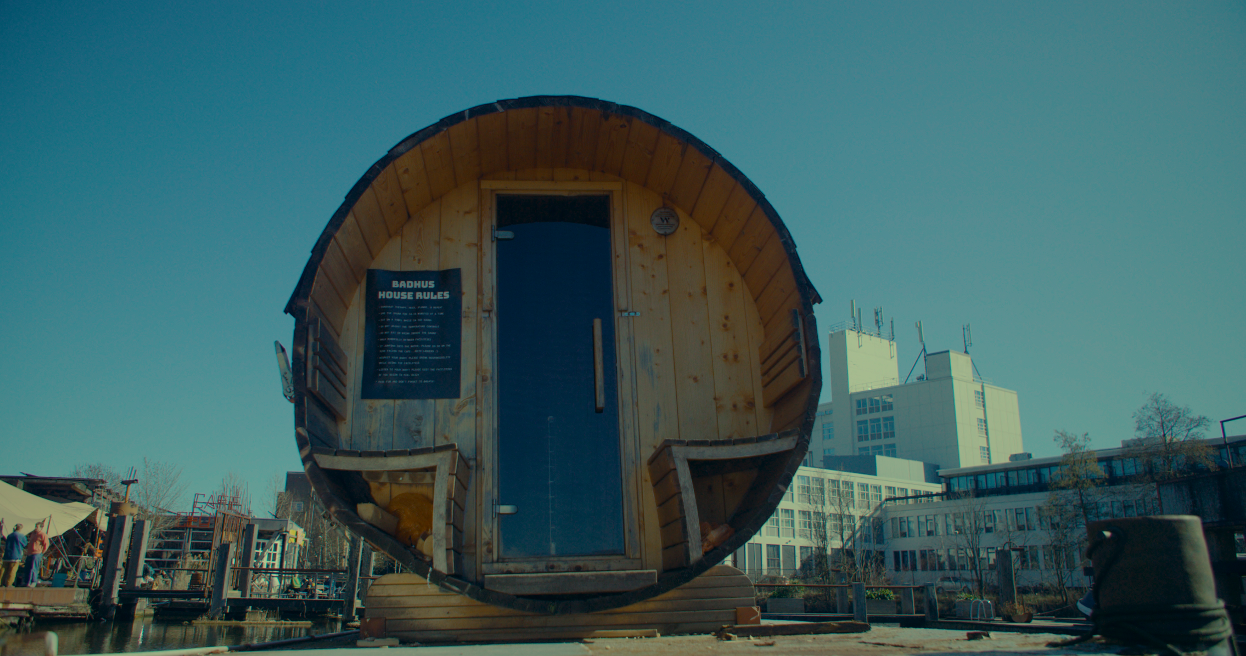 A small round wooden house with a curved roof and a glass door, located outdoors in an urban area, with buildings and trees in the background.