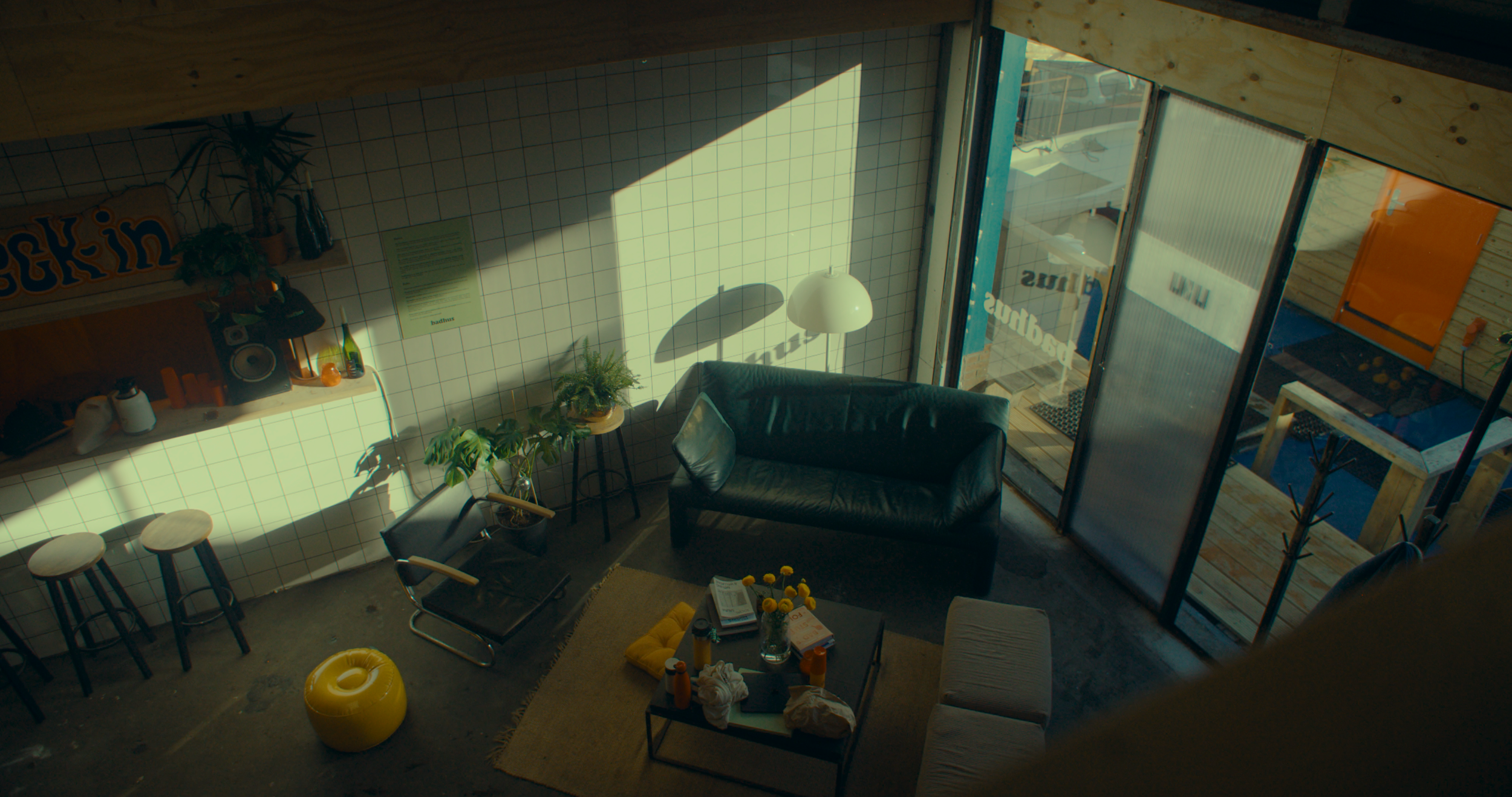 Interior of a cozy living room with a black leather couch, a round glass table with yellow flowers, and green plants near the window. Bright sunlight streams in through large glass doors leading to an outdoor patio.