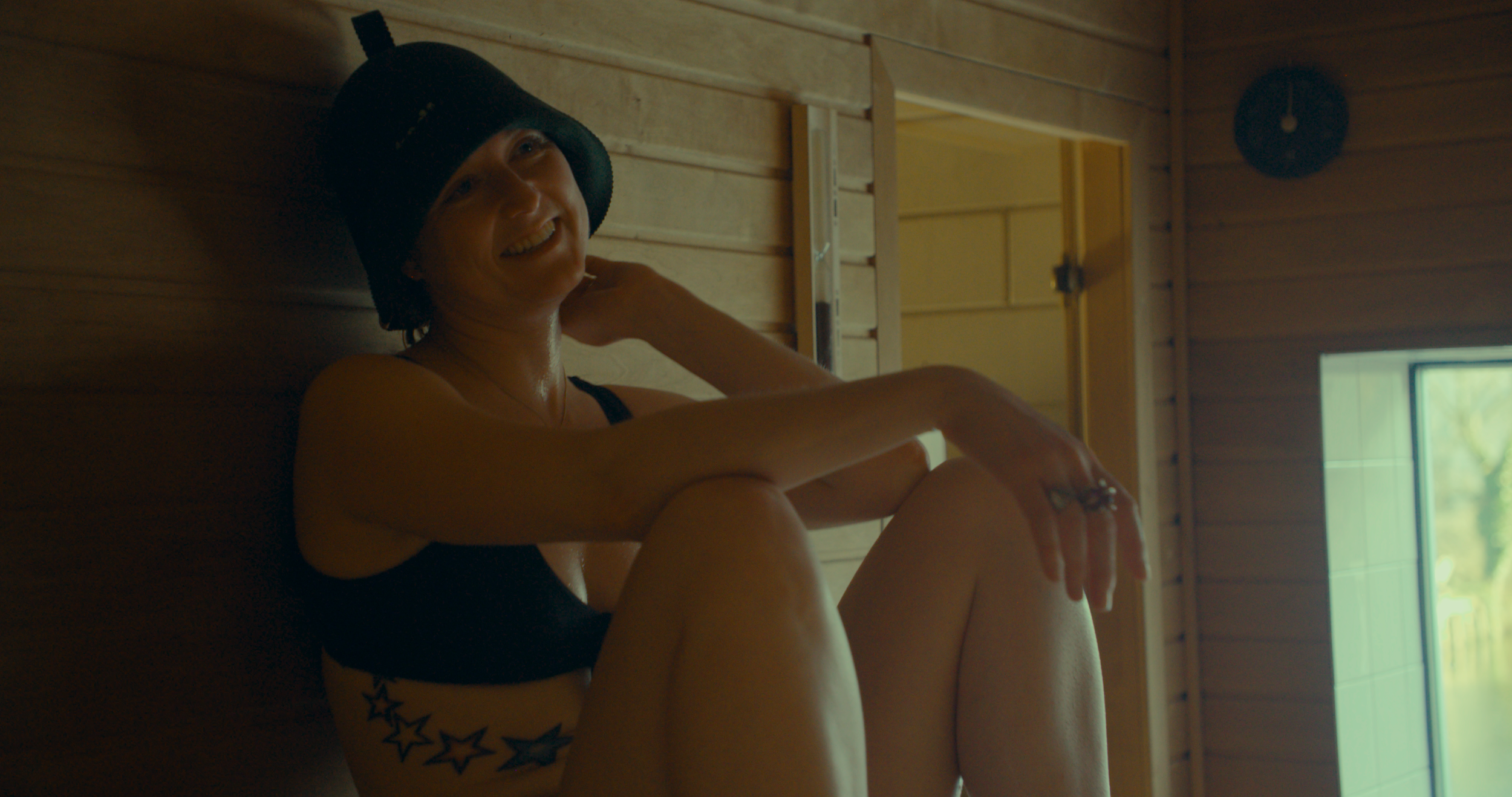 A smiling woman with a star tattoo on her side, wearing a black swimsuit and a black shower cap, sitting in a sauna or steam room with wooden walls and a small window.