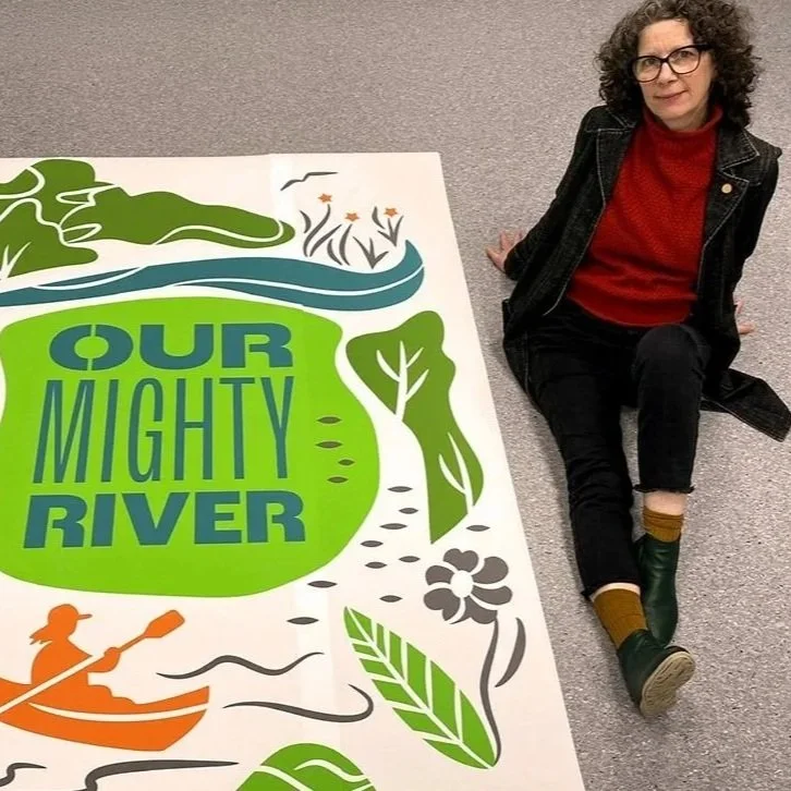 A woman with curly hair wearing glasses, a red sweater, and a black jacket sitting next to a large, colorful sign that reads 'Our Mighty River'. The sign features illustrations of a river, trees, a person kayaking, and flowers in green, blue, orange, and gray colors.