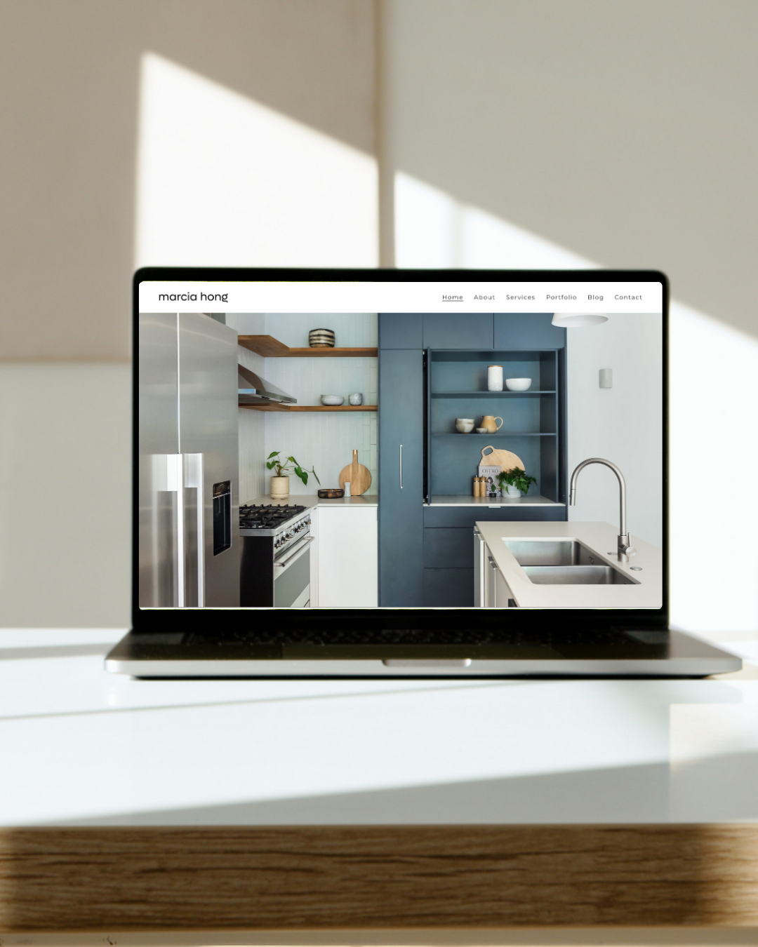 Open laptop on a white table displaying a modern kitchen with blue and white cabinets, wooden shelves, and stainless steel appliances, illuminated by natural light.