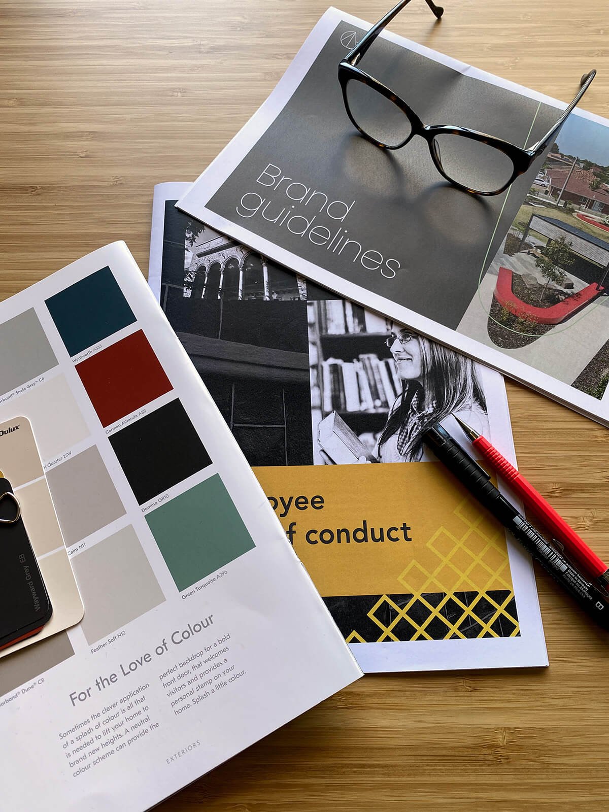 A wooden desk with a pair of black framed glasses resting on a magazine titled "Brand guidelines," along with color swatches, black and white photographs of a woman reading, a pen, and a yellow booklet titled "Employee code of conduct."