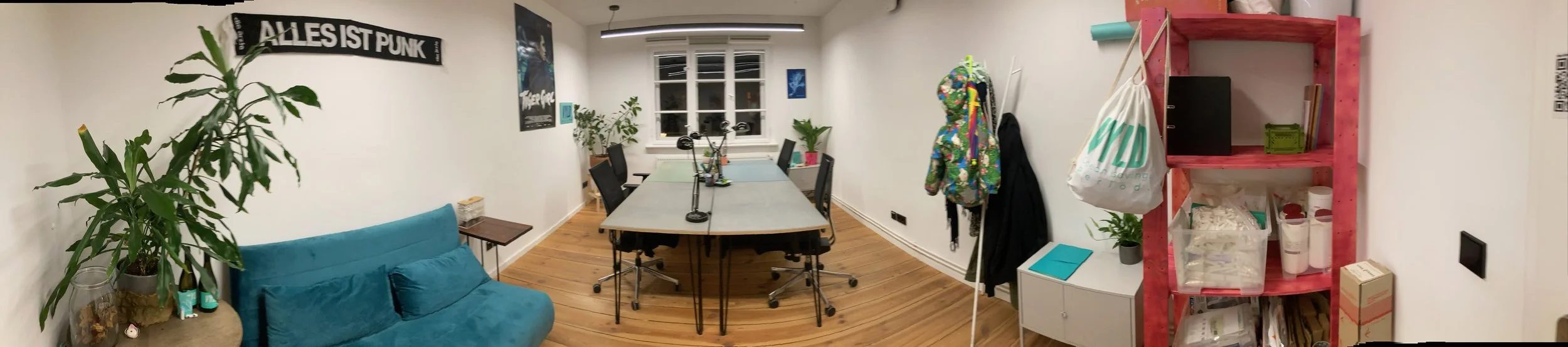 A meeting room with a large central table surrounded by six chairs, decorated with plants and posters, with a red shelving unit on the right, a couch on the left, and a window at the back.