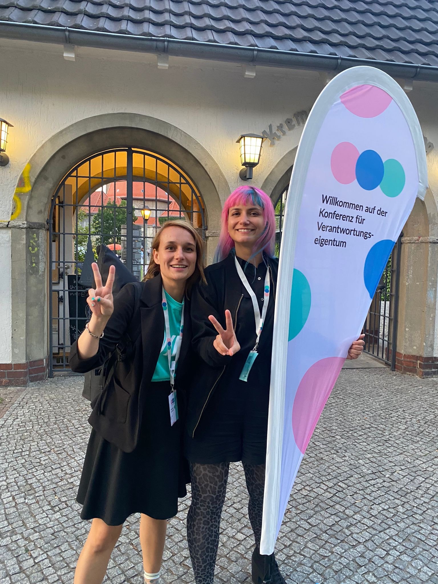 Two women smiling and posing for the camera outside a building, holding a colorful banner with German text, in front of a gated entrance with arched windows and brick details.