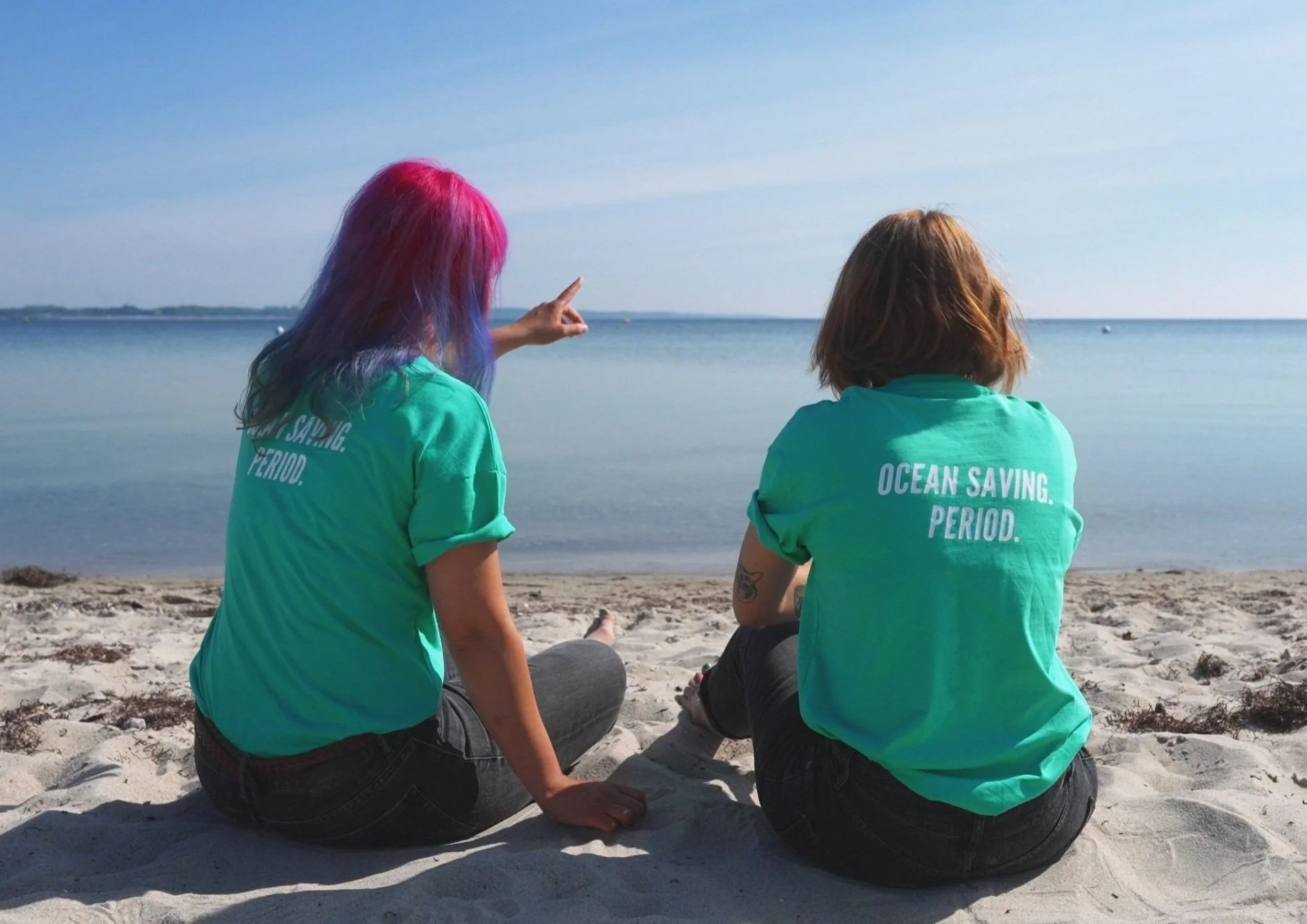 Two women sitting on a sandy beach, facing the ocean. One woman with colorful pink and purple hair is pointing towards the water, both are wearing turquoise t-shirts that say 'OCEAN SAVING. PERIOD.'