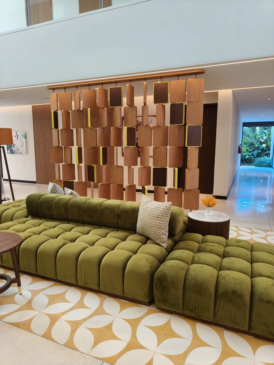 Modern hotel lobby with green velvet tufted sofa, patterned pillows, a side table with orange decorative item, and a wooden room divider with geometric cutouts, overlooking a hallway and outdoor greenery.