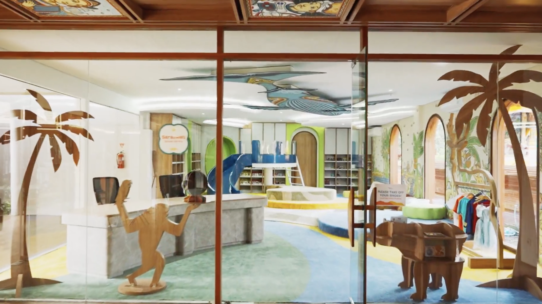 Indoor children's play area with jungle-themed decorations, a slide, and play mats, viewed through glass with palm tree and animal decals.