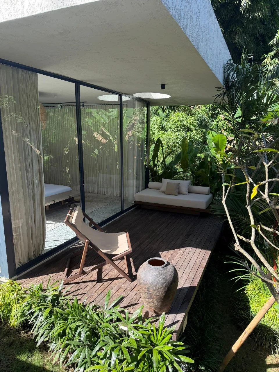 A small outdoor patio enclosed by glass walls, furnished with a reclining lounge chair, a white cushioned daybed, and a large ceramic vase, surrounded by lush green tropical plants.