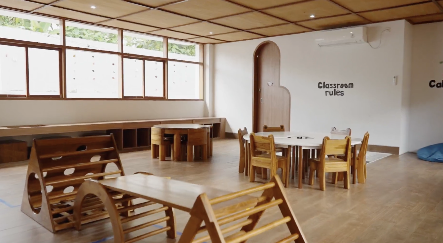 A classroom with wooden tables and chairs, large windows, and sign reading 'Classroom Rules' on the wall.