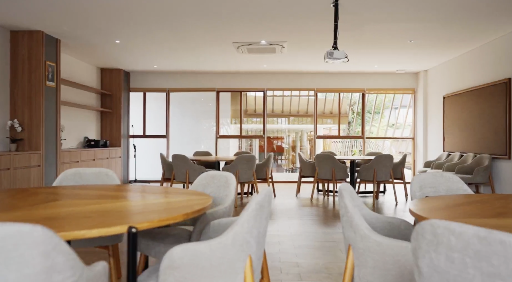 Empty dining room with wooden tables and beige chairs, large windows with wooden frames, and a projector mounted on the ceiling.
