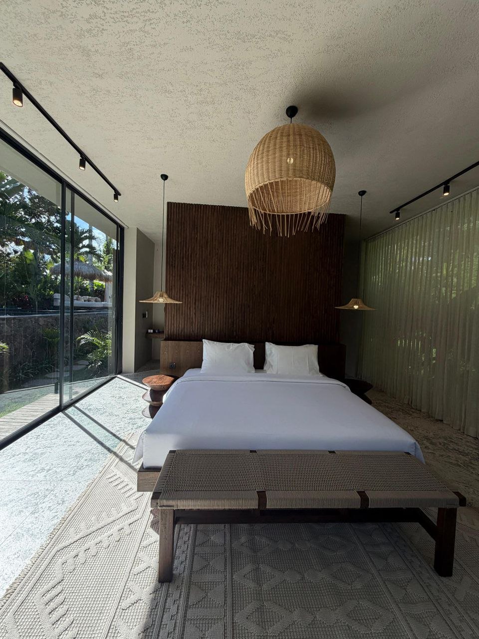 Modern bedroom with a large bed, white linens, two pillows, wooden headboard, hanging wicker light fixture, glass sliding doors, natural light, and a view of greenery outside.