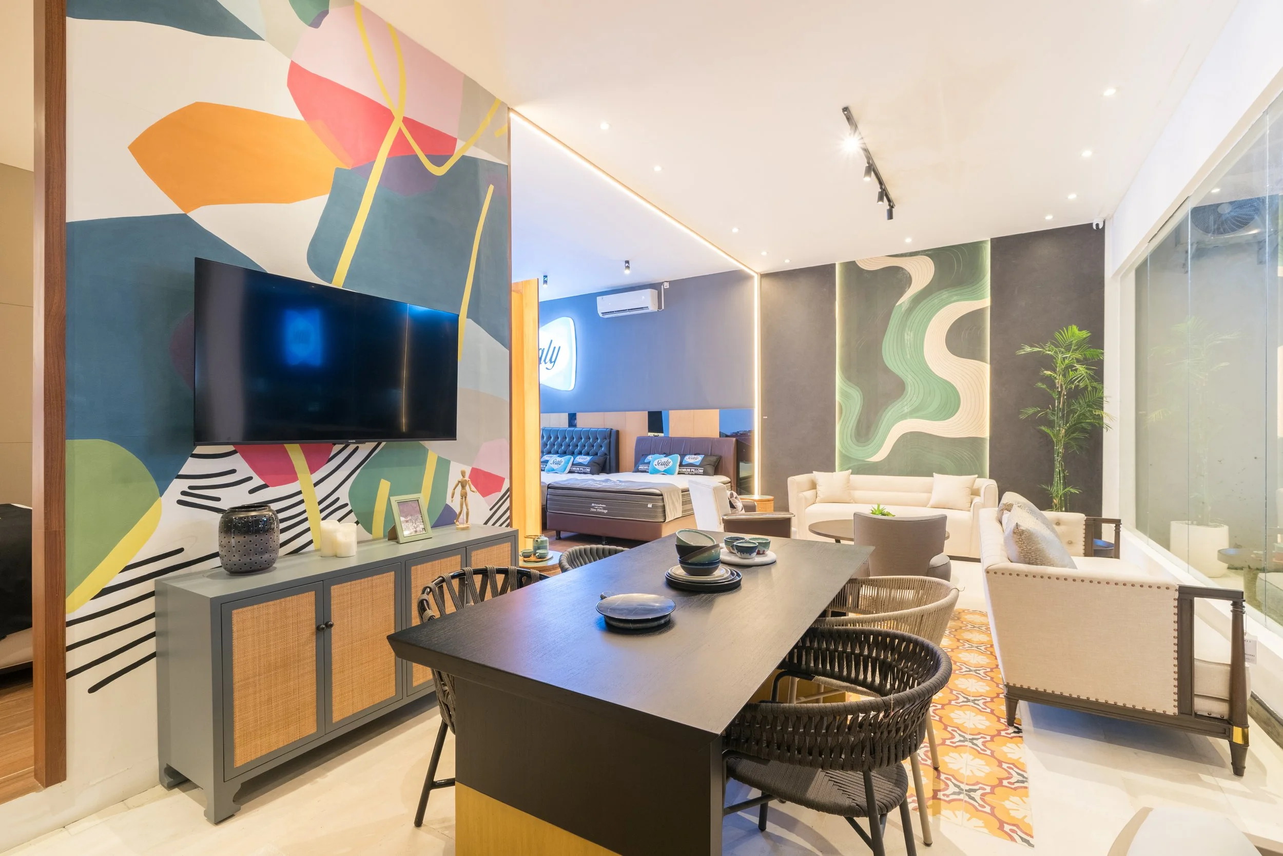 Modern living room with colorful abstract wall art, a grey sideboard, a black dining table with woven chairs, and a seating area with cream sofas, a patterned rug, and decorative elements.