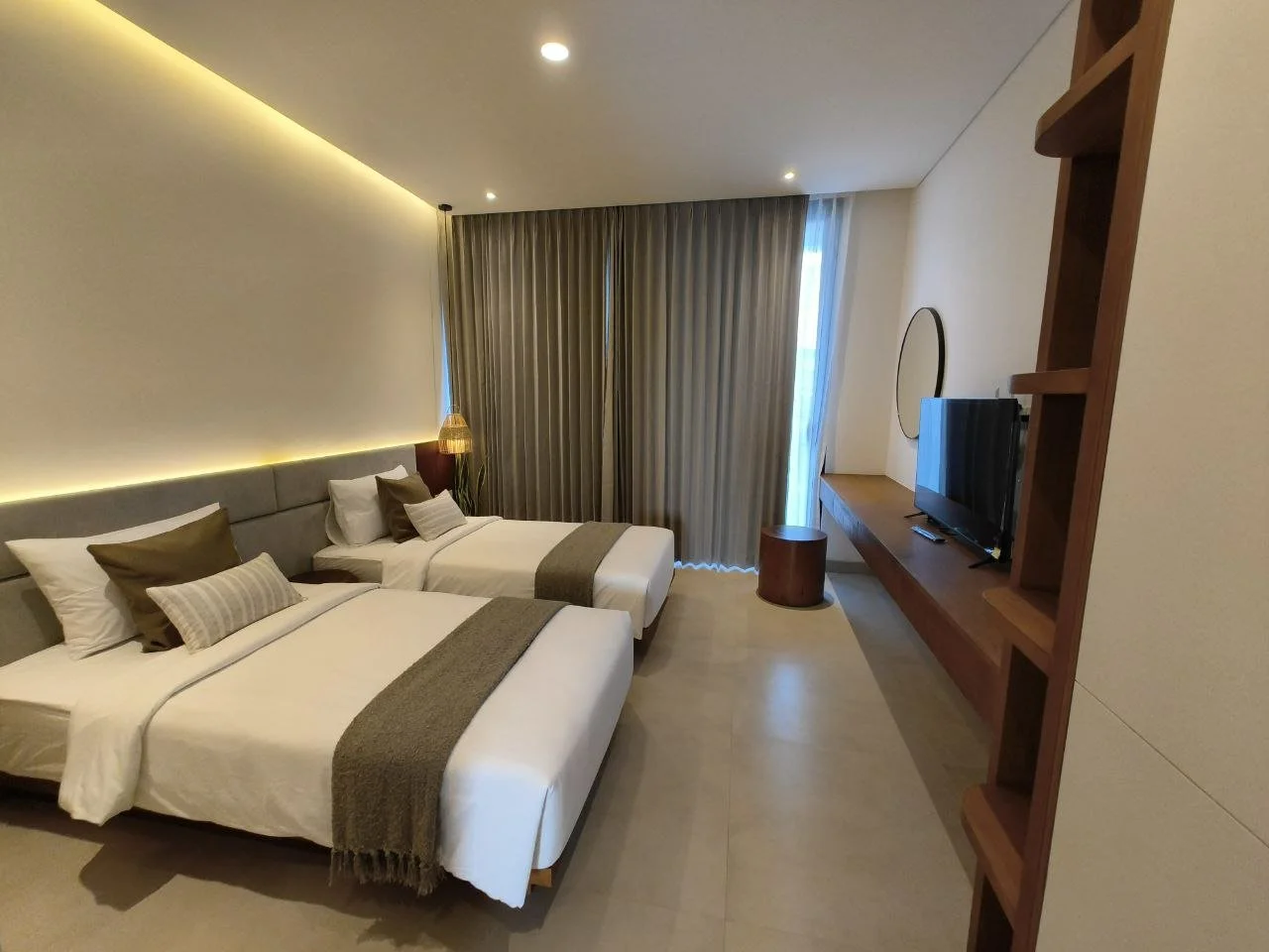 A modern hotel room with two beds, each with white linens, gray headboards, and brown pillows. There is a small bedside table with a lamp between the beds. A large window with beige curtains is at the back. A flat-screen TV is mounted on the wall opp