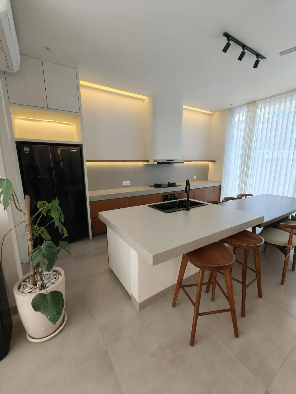 Modern kitchen with white island, black sink and faucet, gray countertops, wooden cabinets, black refrigerator, spot lighting, large window with sheer curtains, potted plant, and wooden bar stools.