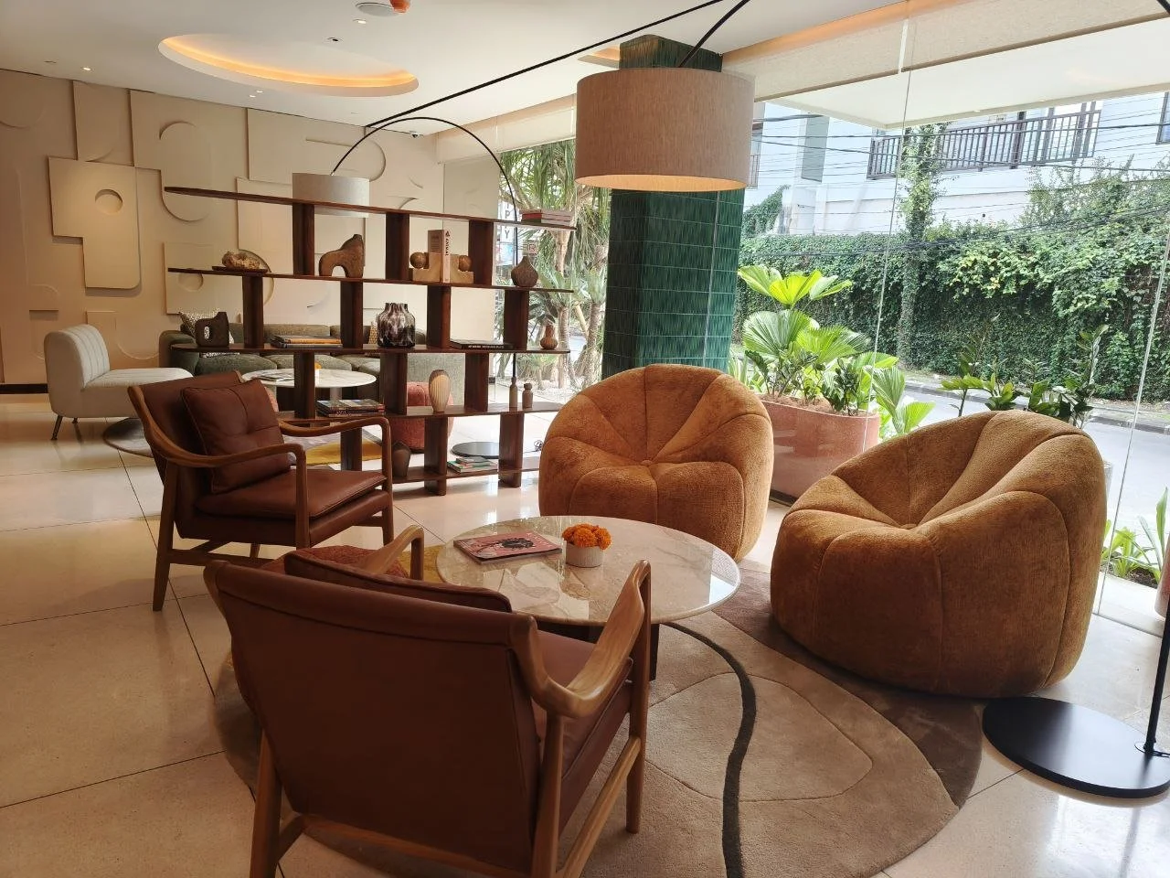 Modern hotel lobby with beige and green decor, featuring comfortable armchairs, a marble coffee table with flowers, shelves with decorative items, and large windows with plants outside.