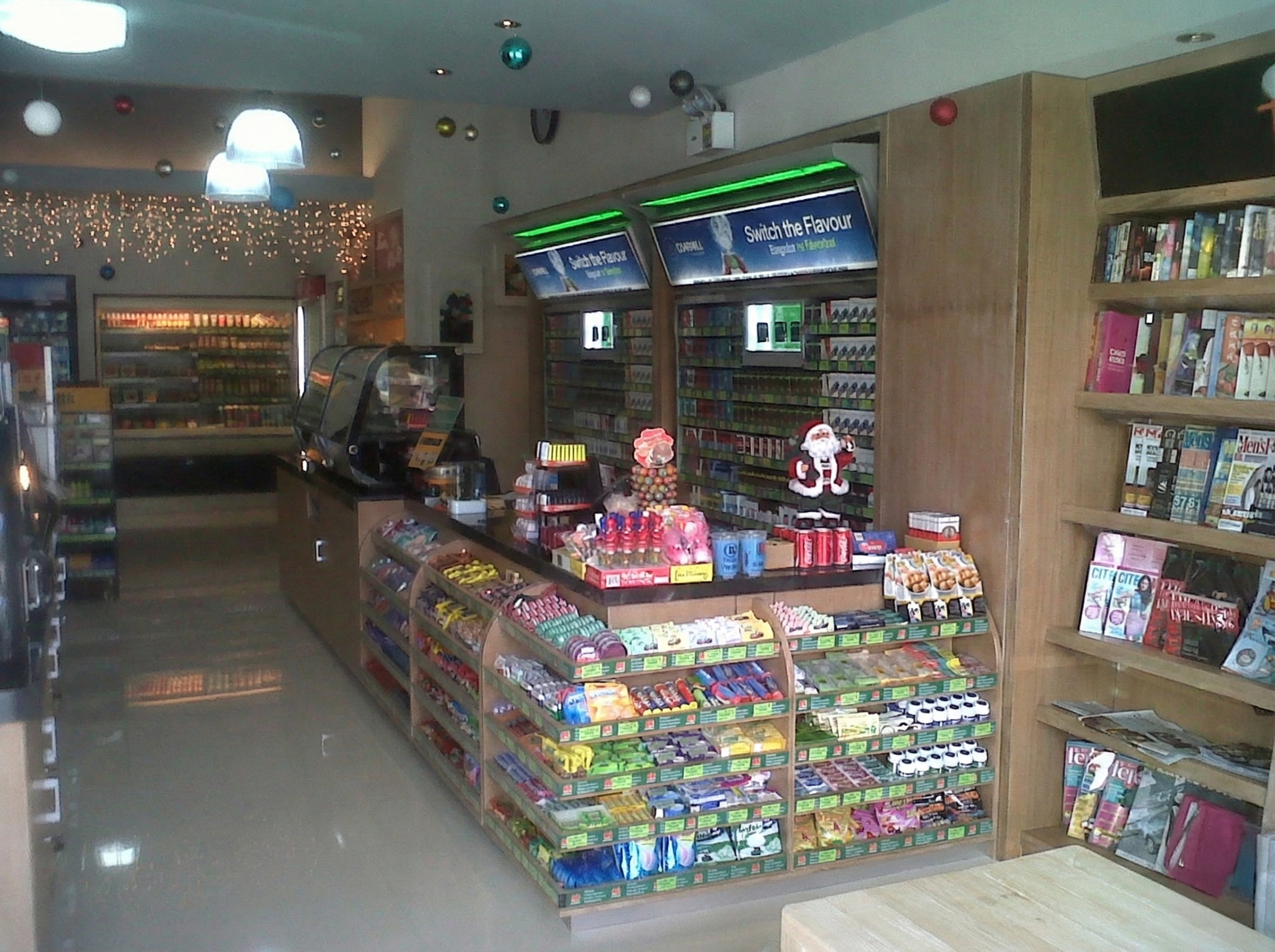 Inside a convenience store decorated for Christmas, with shelves stocked with snacks, magazines, and festive decorations including a small Santa figure and Christmas ornaments.