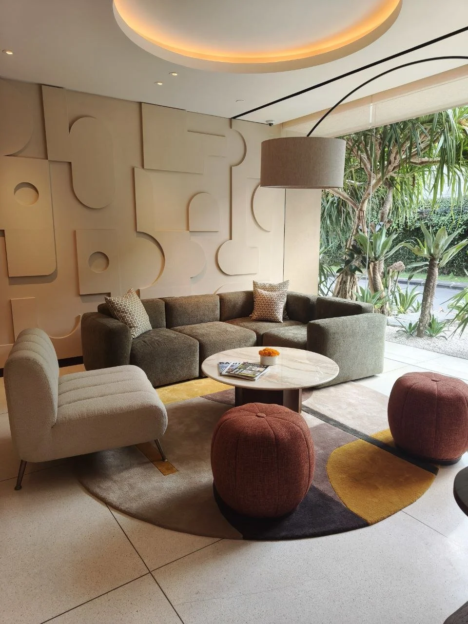 Modern hotel lobby seating area with a gray sectional sofa, a beige armchair, two red ottomans, a round marble coffee table, and decorative pillows. Large window with outdoor greenery visible.