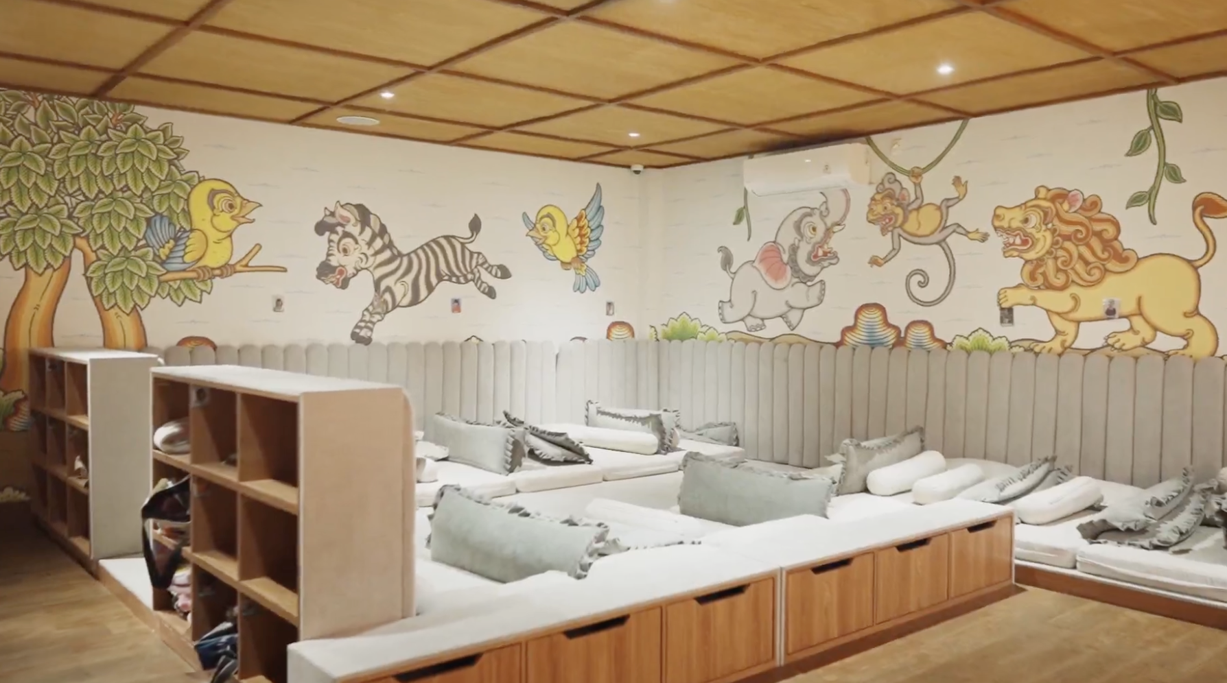 Colorful mural featuring a decorated jungle scene with a bird, zebra, elephant, monkey, and lion, painted on a white wall in a cozy room with beige cushions on low wooden seating.