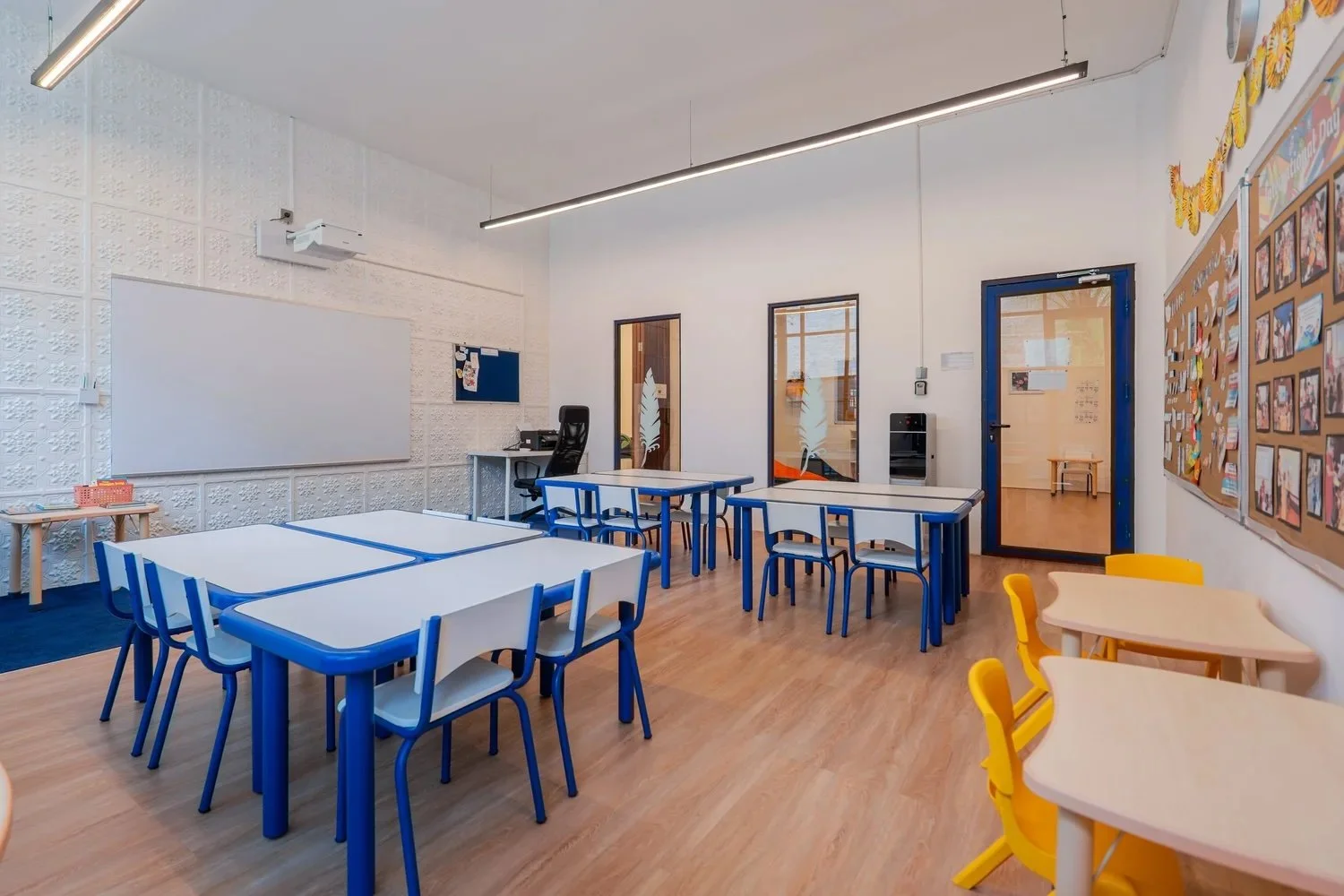 Empty classroom with blue and yellow tables and chairs, whiteboard, bulletin board, door, window, and wooden flooring.