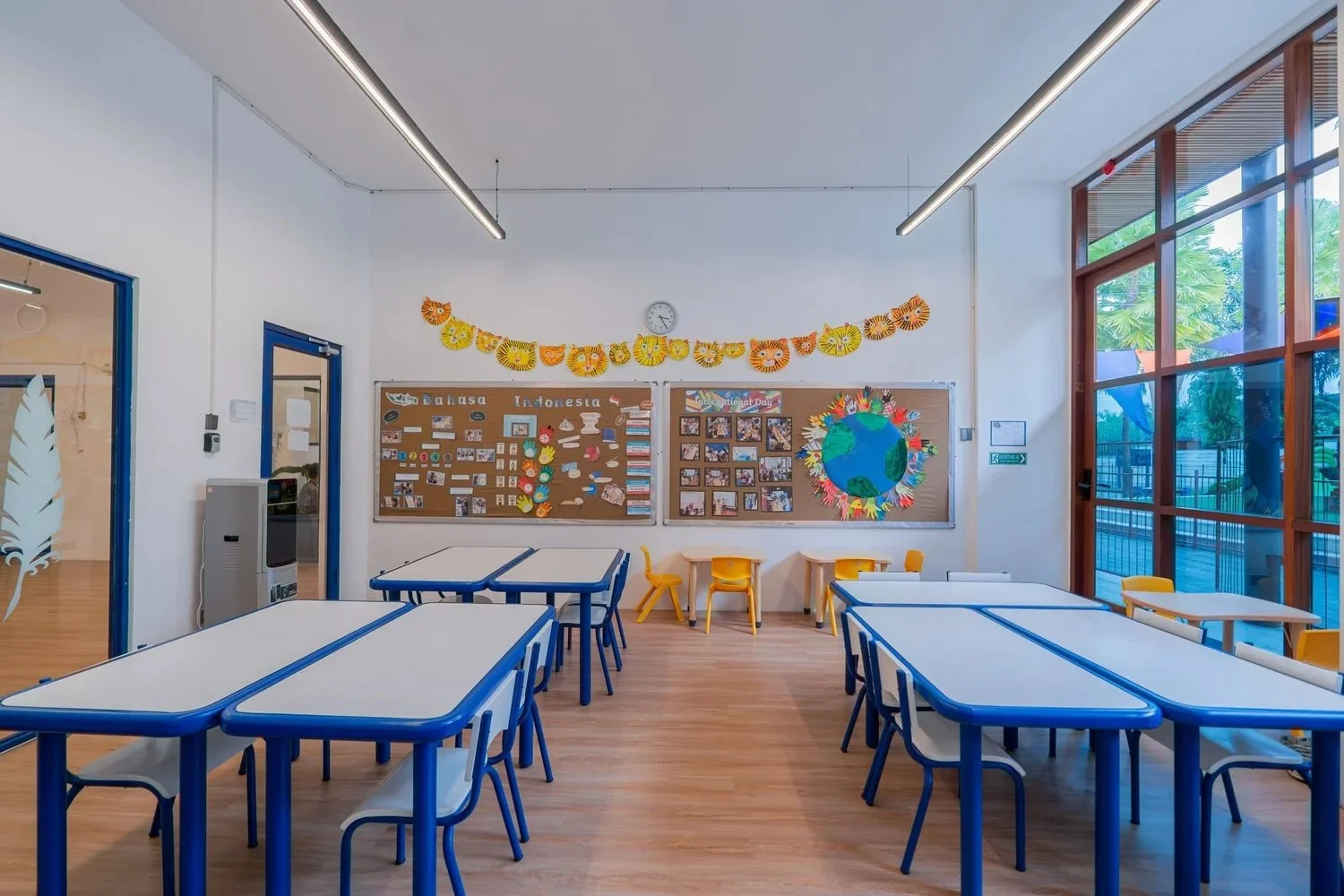 Empty classroom with white tables and yellow and black chairs, decorated bulletin boards about Indonesia, a globe with colorful paper flames, and a large window showing trees outside.