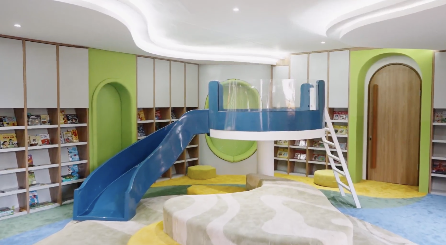 Indoor children's play area featuring a blue slide, a small playhouse with a rounded opening, surrounded by children's bookshelves, green and yellow walls, and colorful carpet floor.