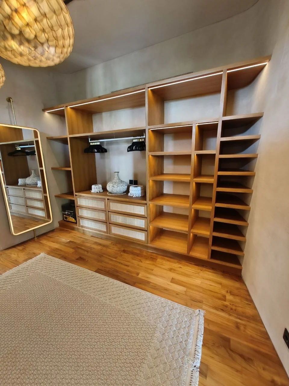 Empty wooden built-in closet with open shelves and hanging space, decorated with vases and a mirror nearby, in a room with hardwood floor and beige rug.