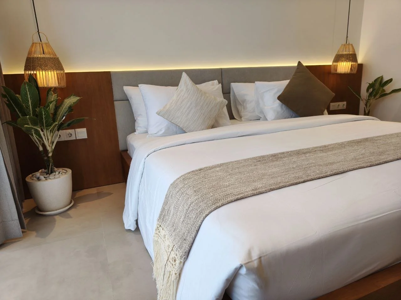 A neatly made bed with white linens, beige and brown throw pillows, and a beige bed runner in a modern hotel room. There are two potted plants and hanging pendant lights on either side of the bed.