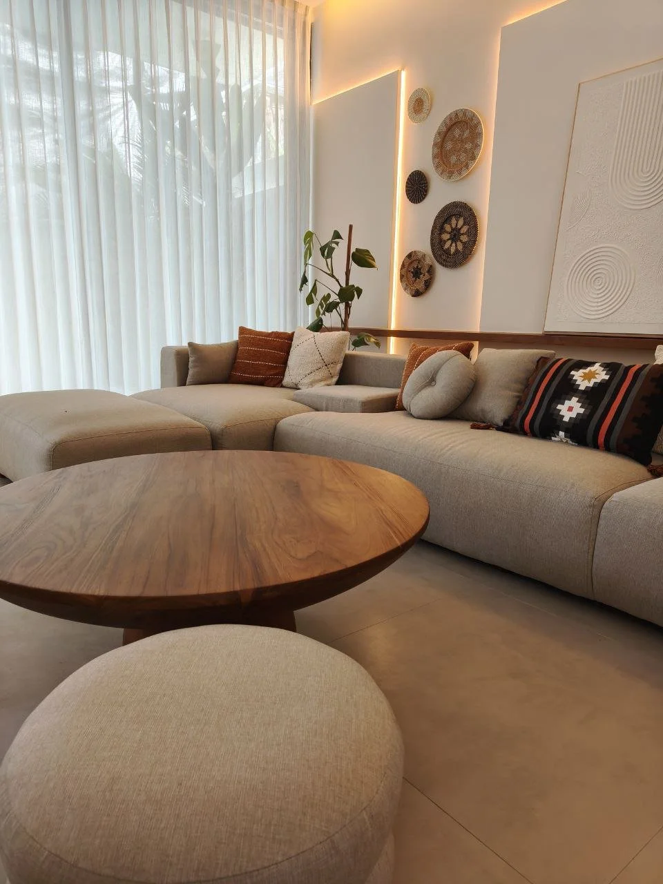 Modern living room with beige sectional sofa, decorative pillows, round wooden coffee table, beige ottoman, sheer curtains, wall art, indoor plant, and soft lighting.