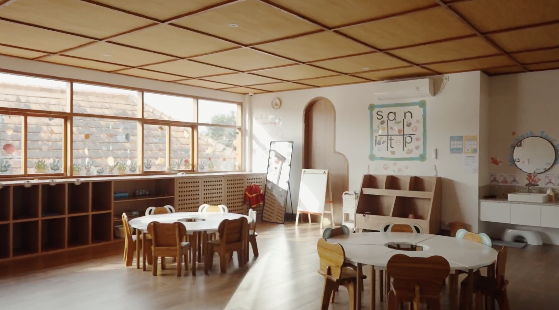 A bright and airy classroom with large windows, wooden ceiling, and small round tables with chairs for children. Decorated with educational posters and art, including a sign with the word "salt" and colorful plants painted on the wall.