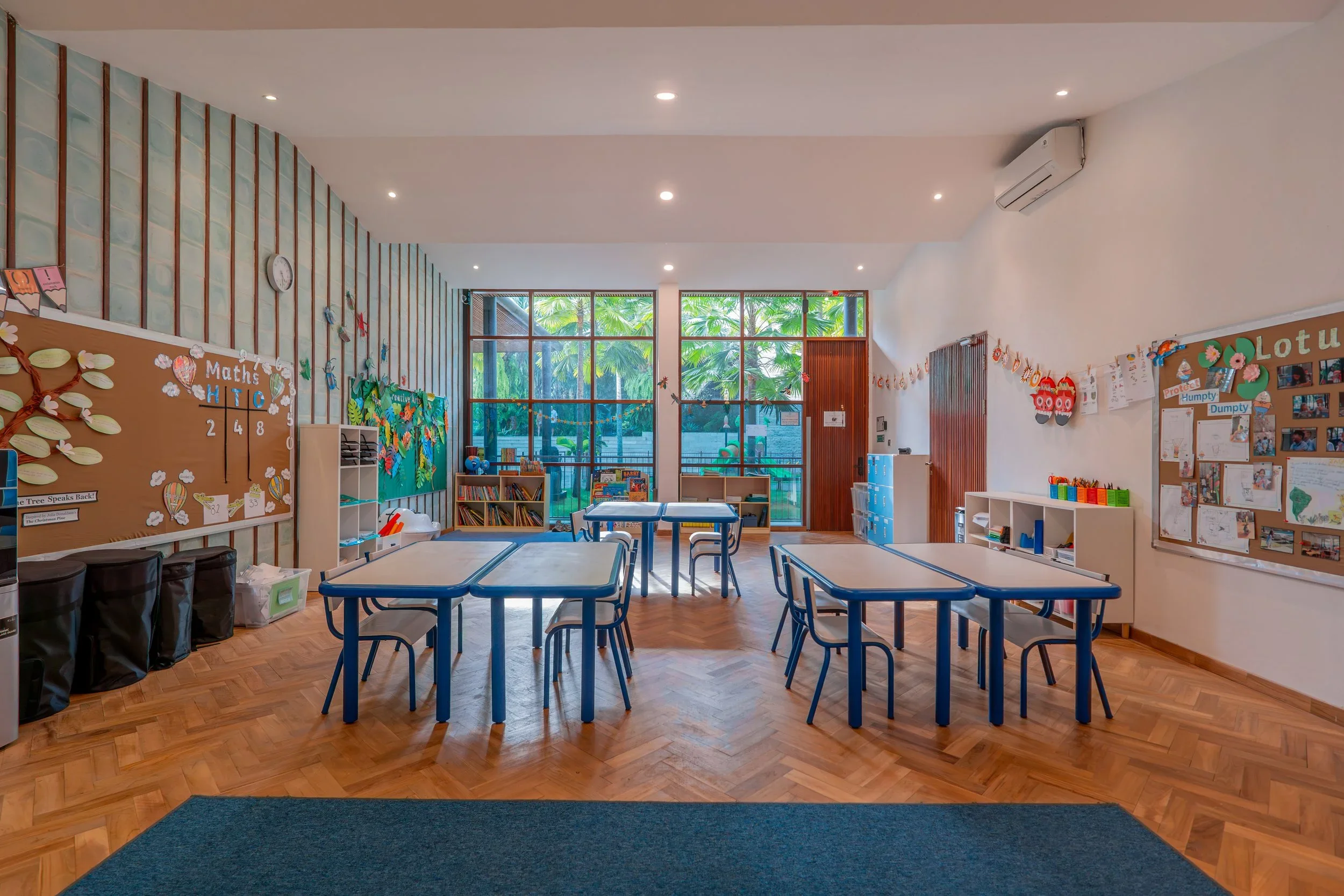 A bright classroom with large windows showing palm trees outside. The room has blue tables and chairs, bulletin boards with educational decorations, bookshelves, and toys for young children.