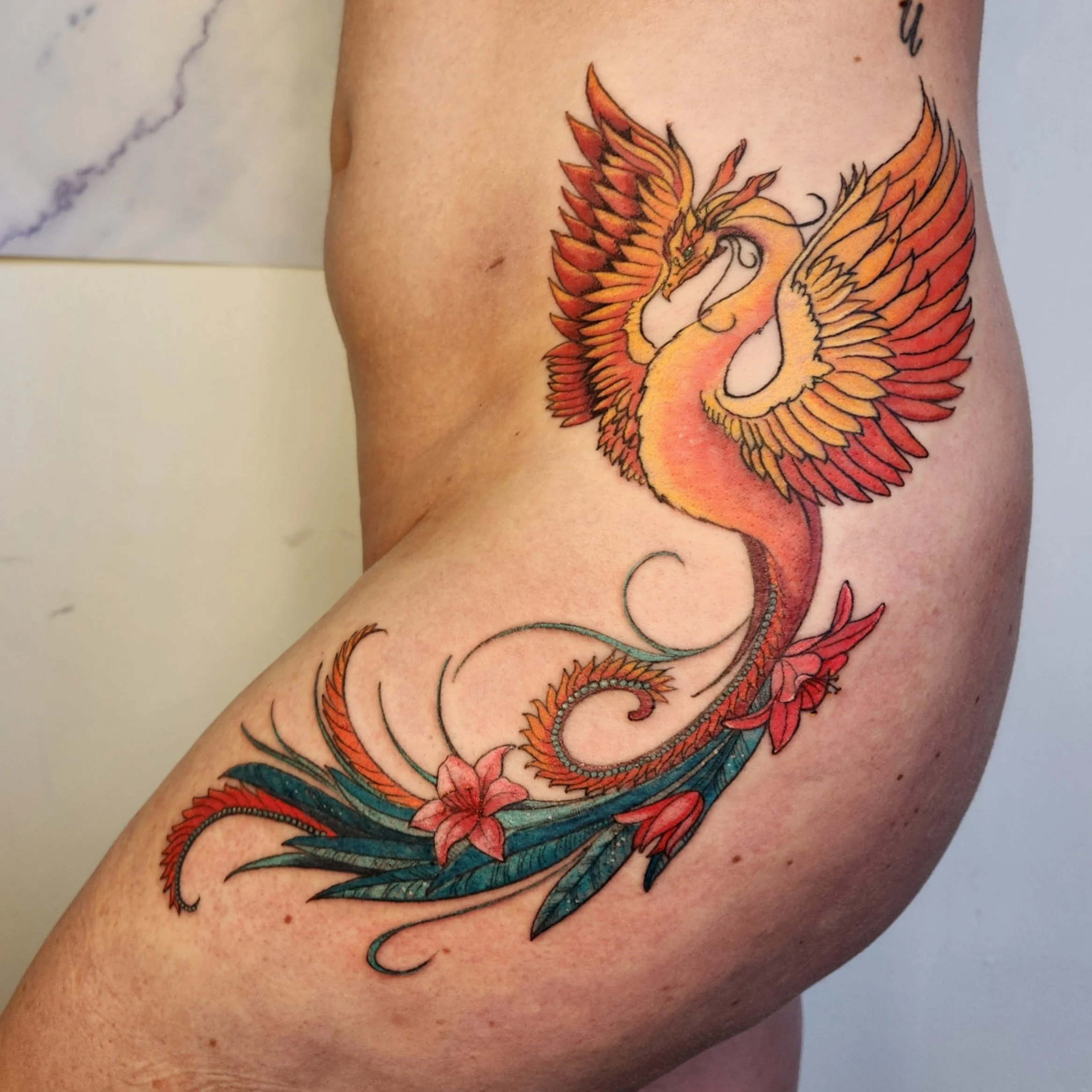 Colorful tattoo of a phoenix with orange, yellow, red, and blue feathers on a person's upper arm and shoulder.