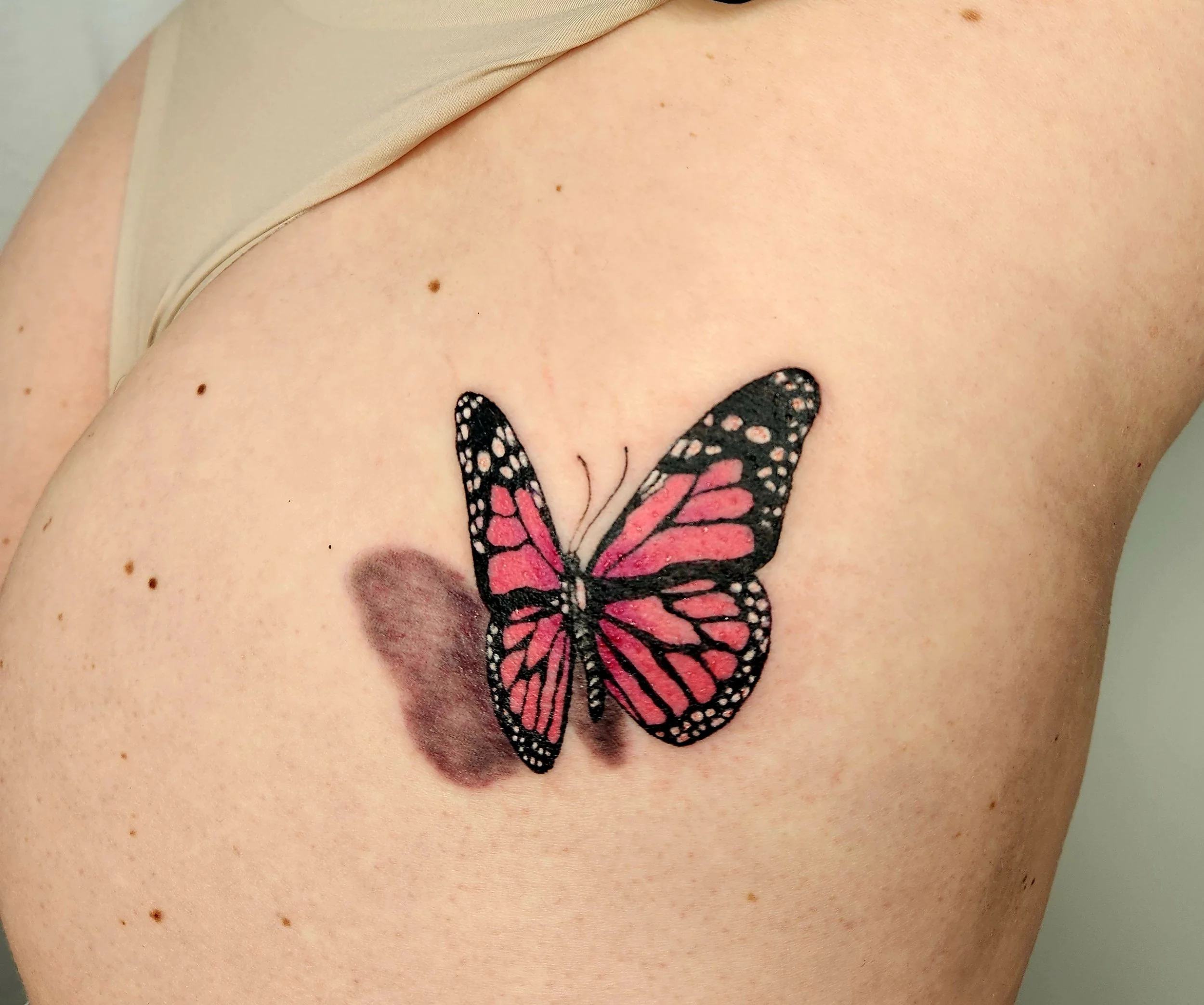 A colorful butterfly tattoo on a person's upper arm, with pink and black wings and shadow details.