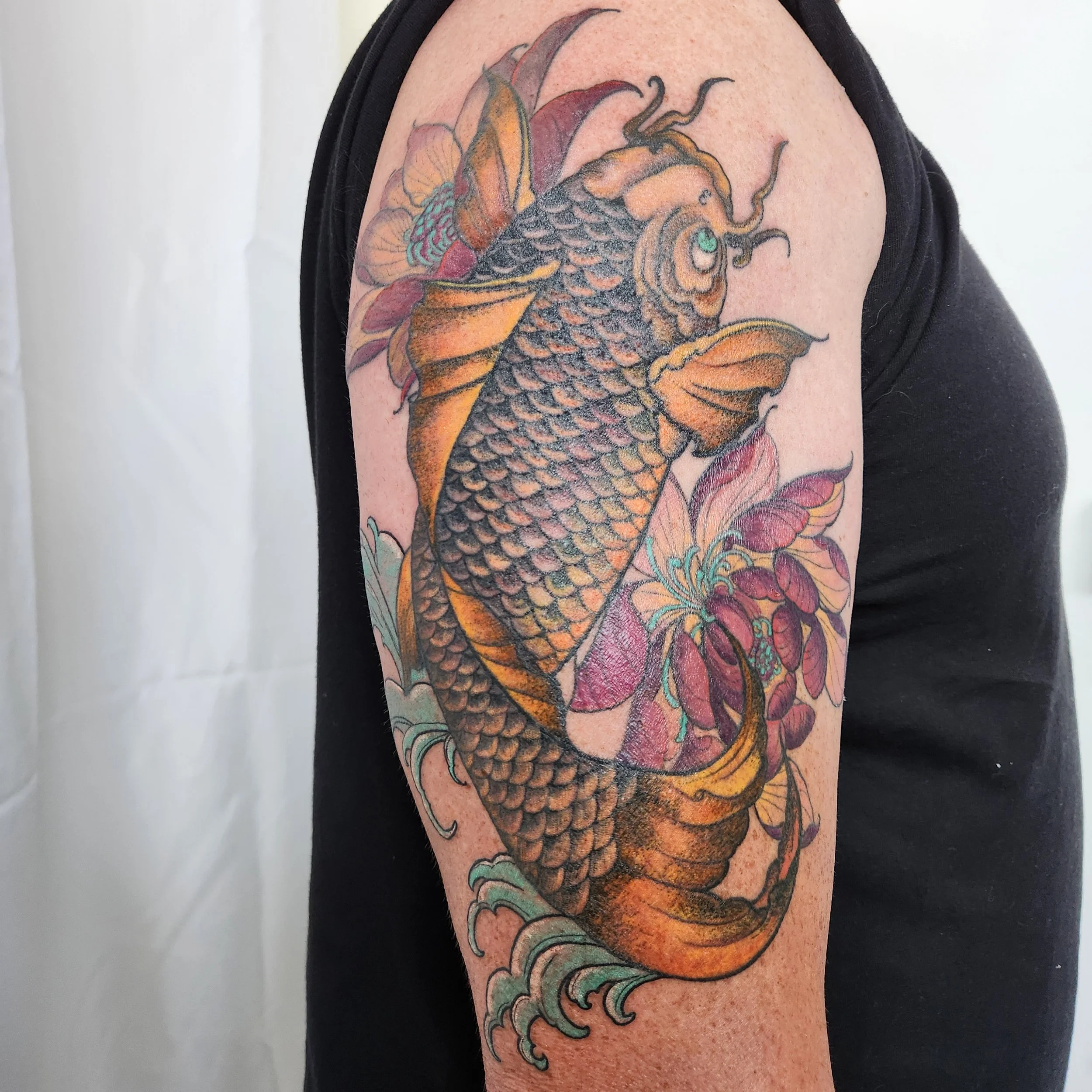 A tattoo of a traditional style koi fish with orange and black scales, surrounded by colorful flowers and water waves on a person's upper arm.