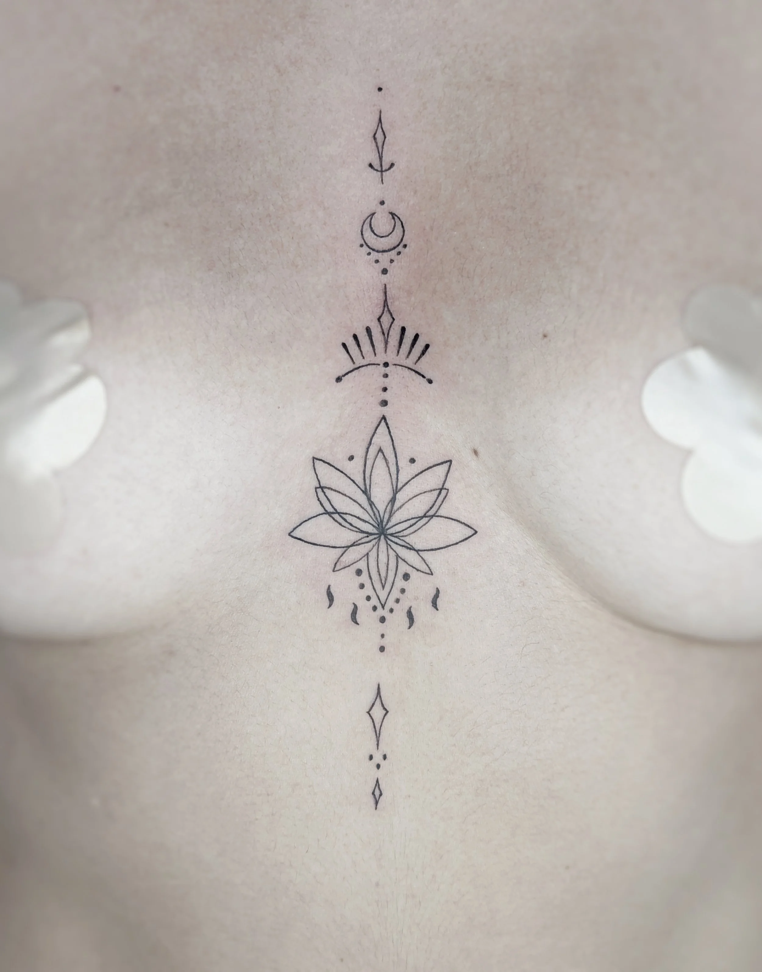 A black line tattoo design on a person's skin featuring a symmetrical floral pattern with geometric elements, crescent moons, and small decorative symbols arranged vertically.
