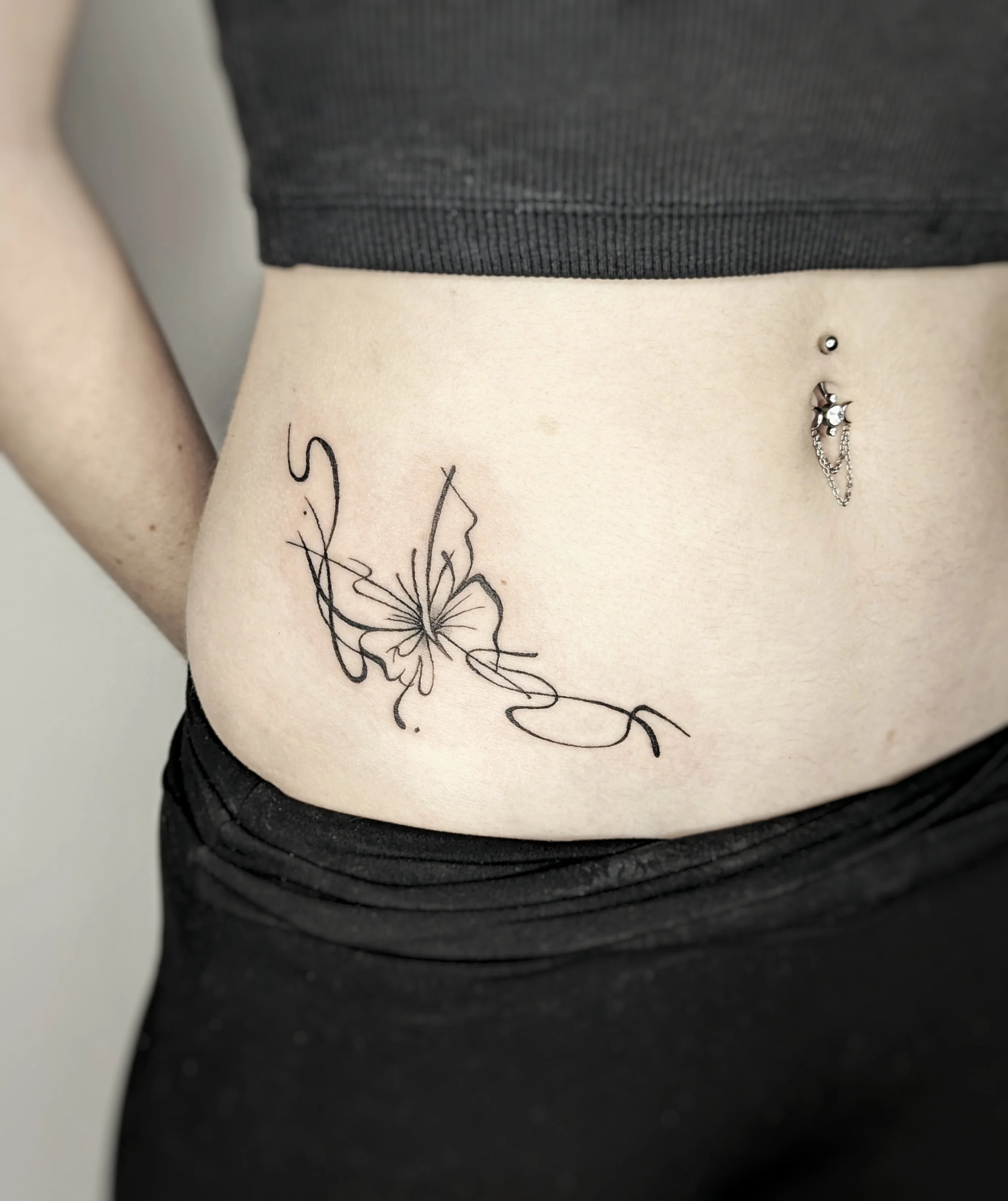 Close-up of a person's side abdomen with a black butterfly tattoo and a belly button piercing with chains and small charms.
