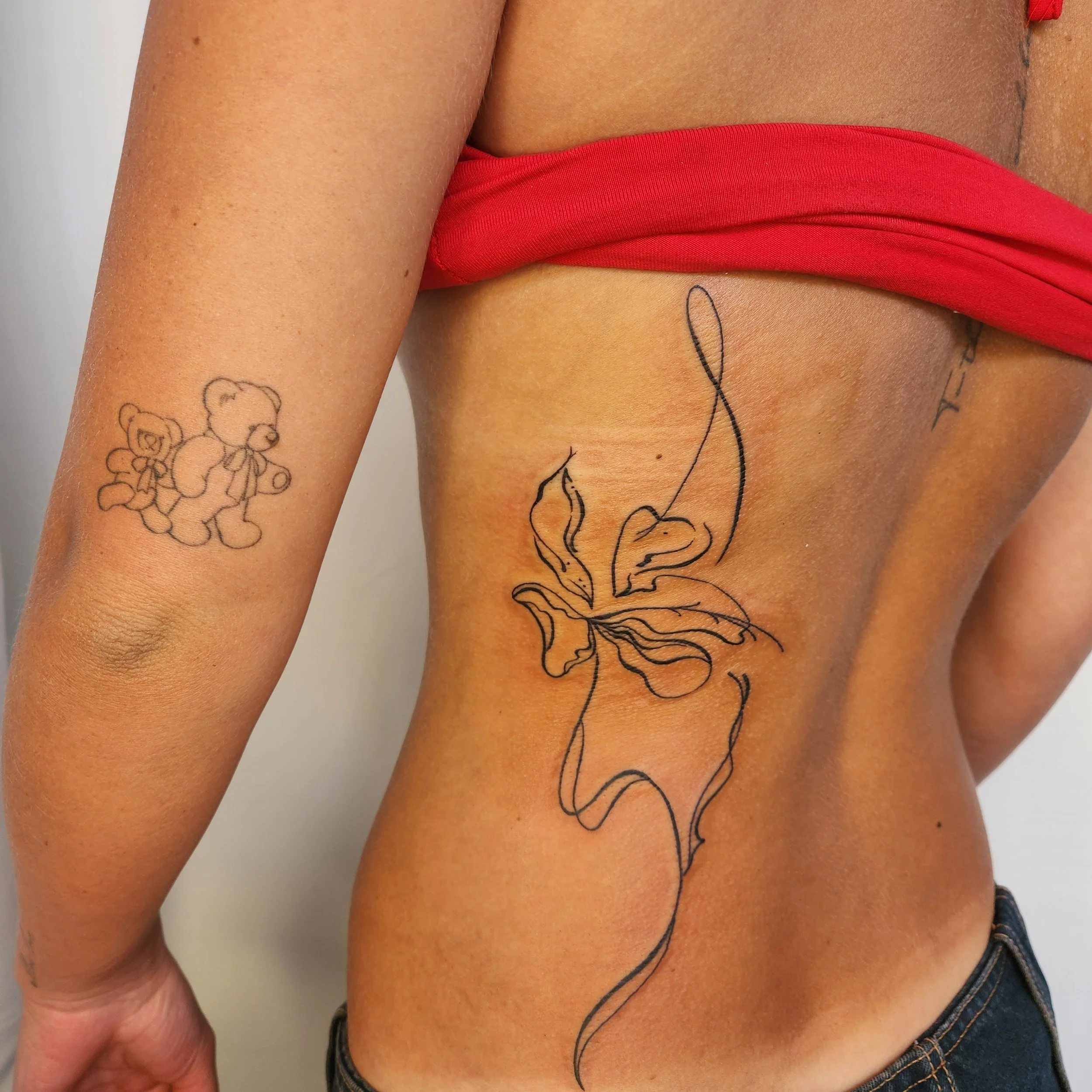 Close-up of a person's side showing two tattoos: a line drawing of a butterfly on their side and a small teddy bear tattoo on their inner arm.